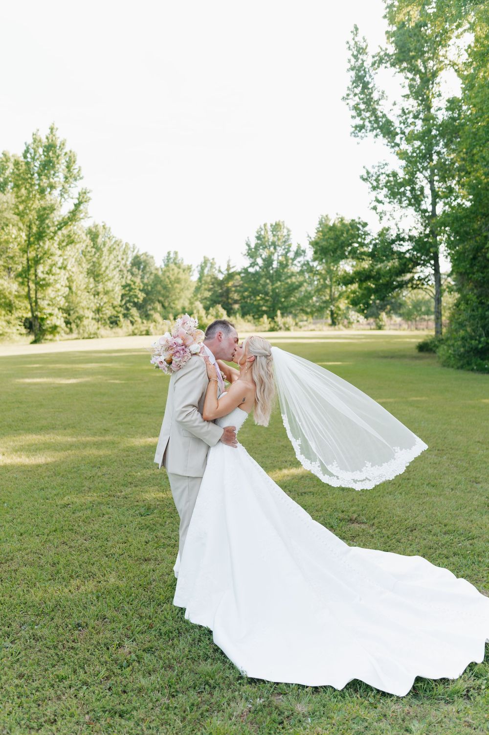 Bride and groom share a romantic kiss on lush green lawn, her white gown and veil flowing gracefully in the breeze.