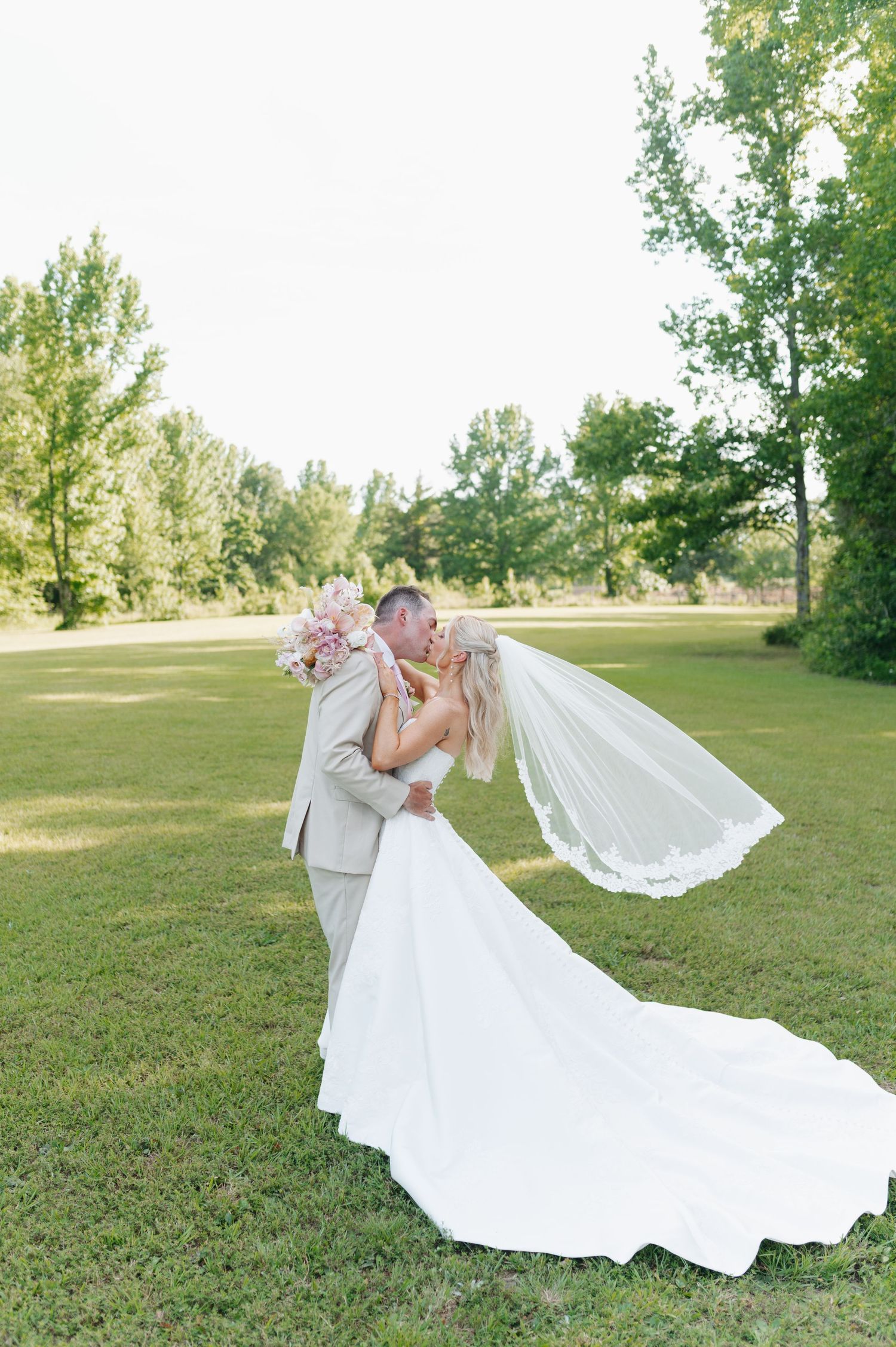Bride and groom share a romantic kiss on lush green lawn, her white gown and veil flowing gracefully in the breeze.