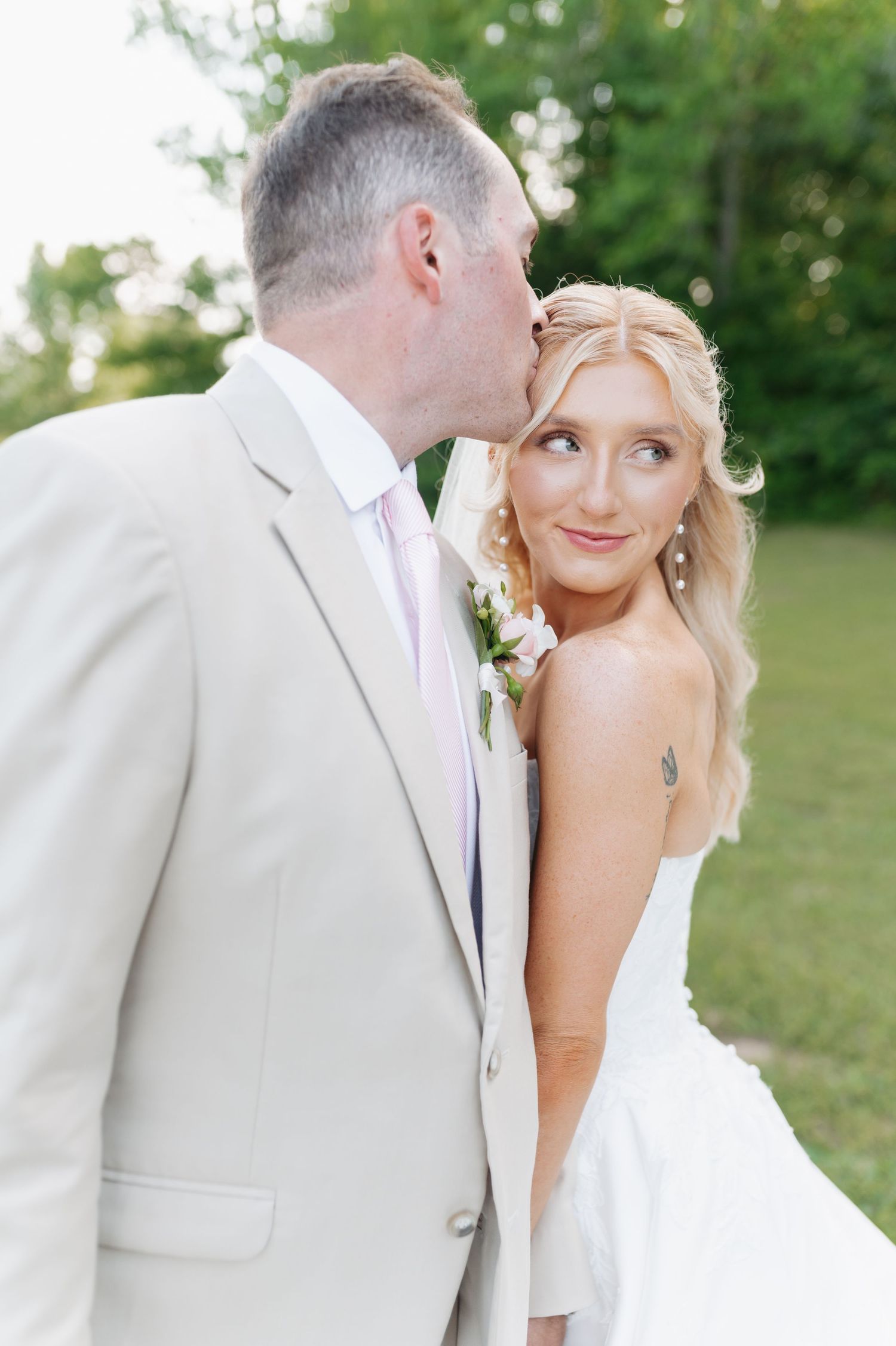 Groom in tan suit kisses bride on cheek as she smiles outdoors at their wedding ceremony.