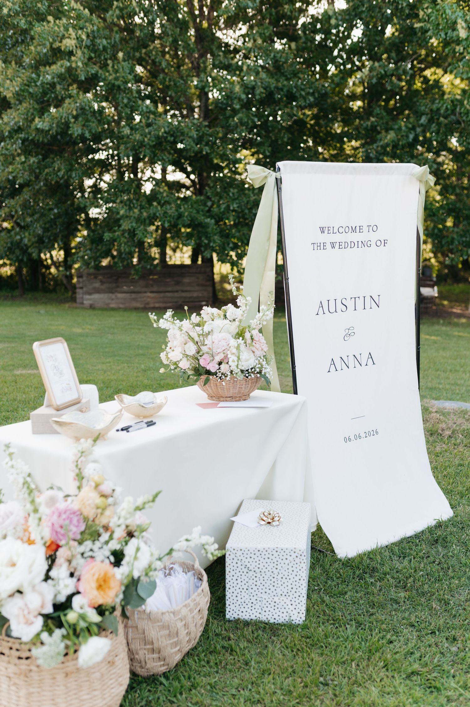 Outdoor wedding welcome sign for Austin and Anna with floral arrangements on a white draped table in a garden setting.