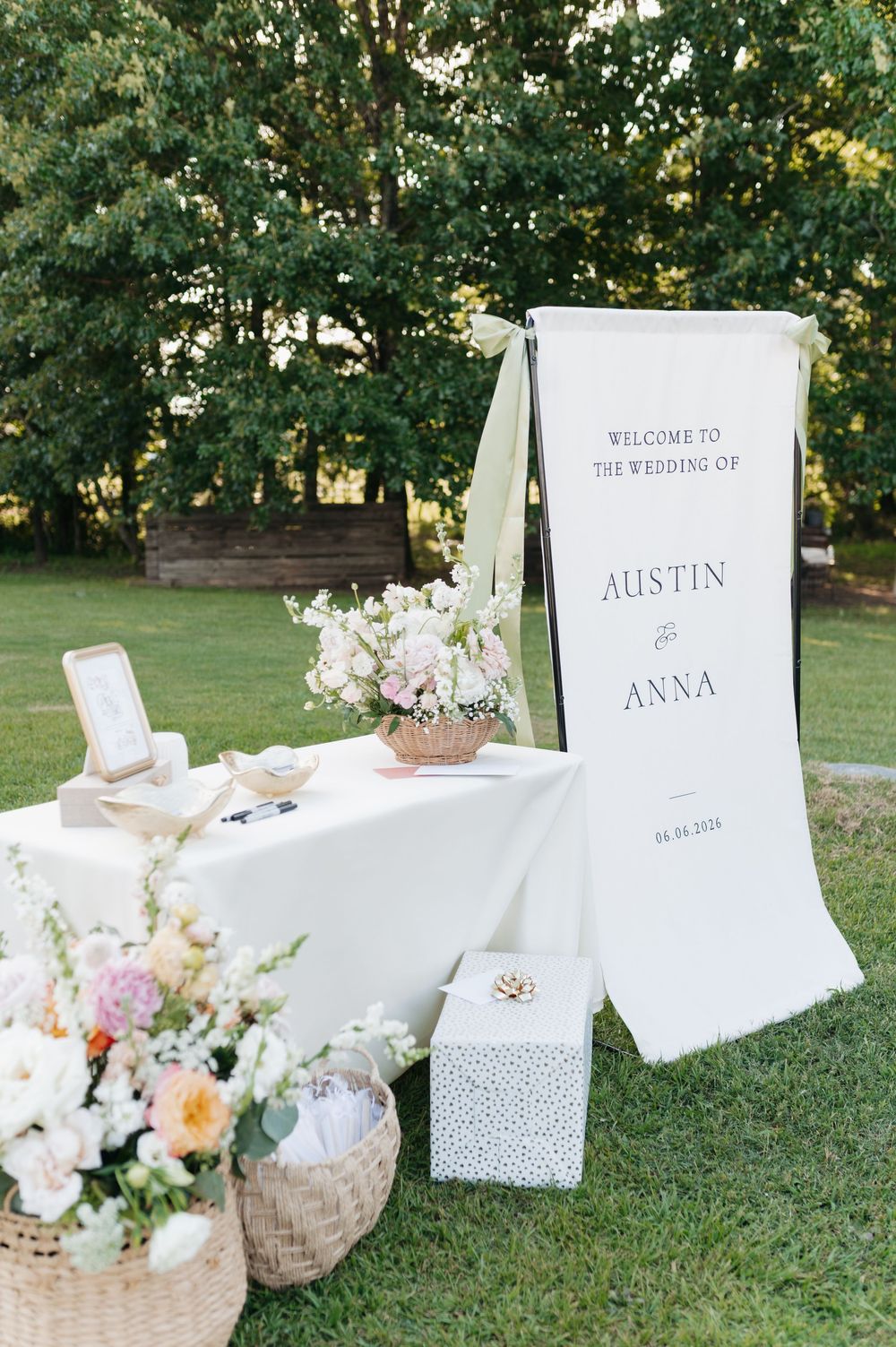 Outdoor wedding welcome sign for Austin and Anna with floral arrangements on a white draped table in a garden setting.