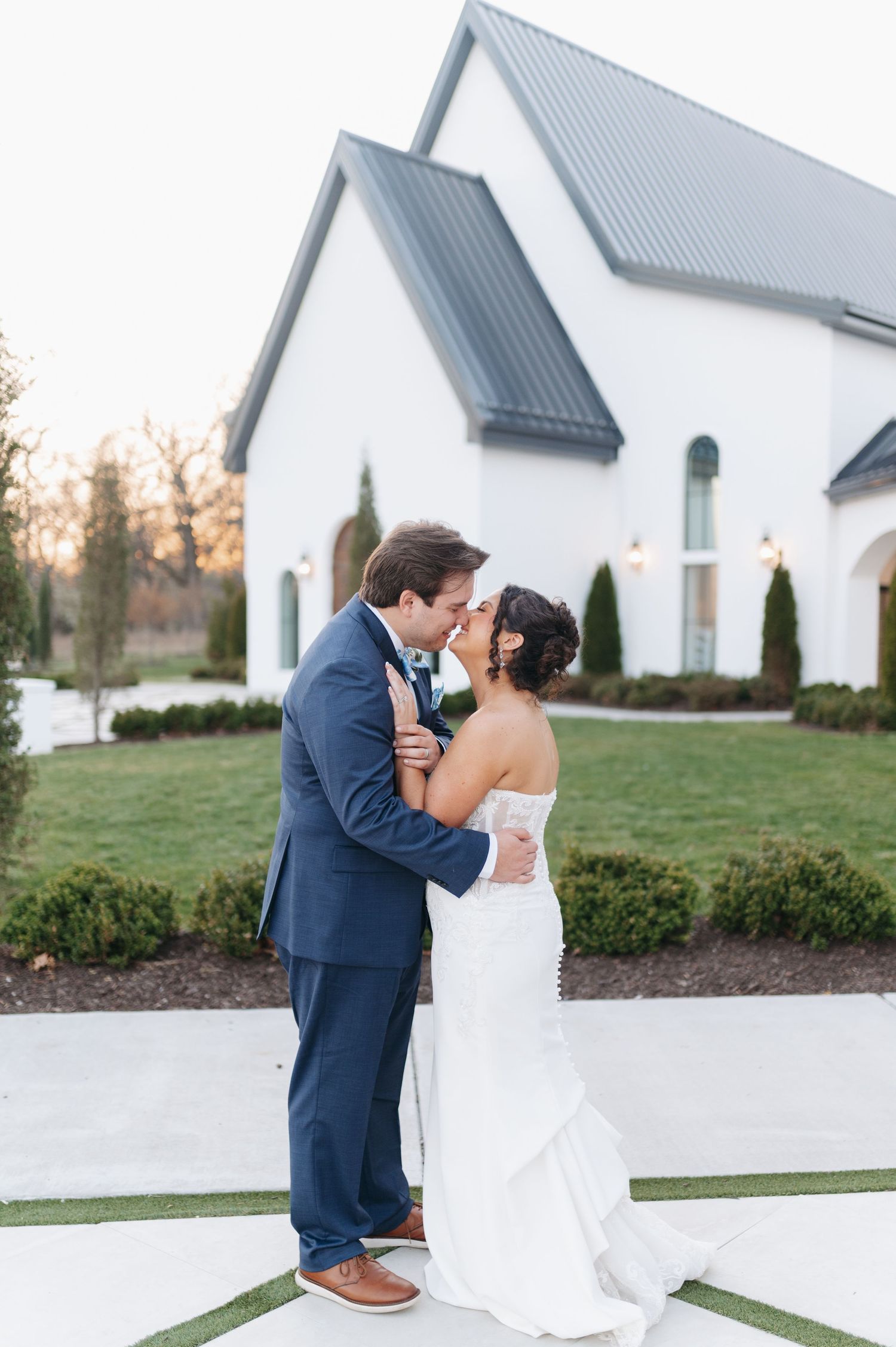 Bride and groom share a kiss outside a white chapel wedding venue surrounded by manicured gardens.
