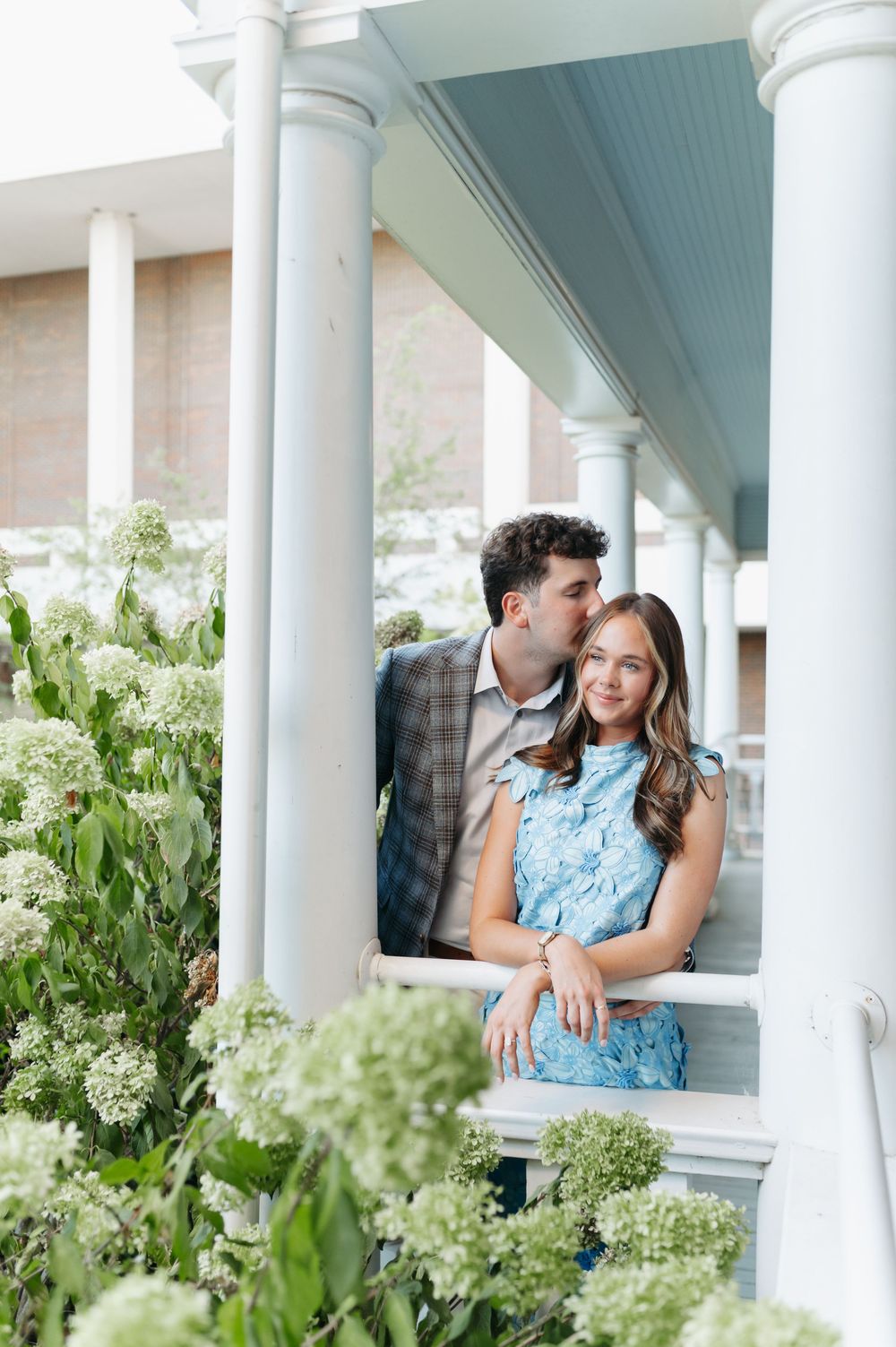 Couple poses on a porch surrounded by white hydrangeas, man kissing woman's cheek as she smiles in a blue lace dress.
