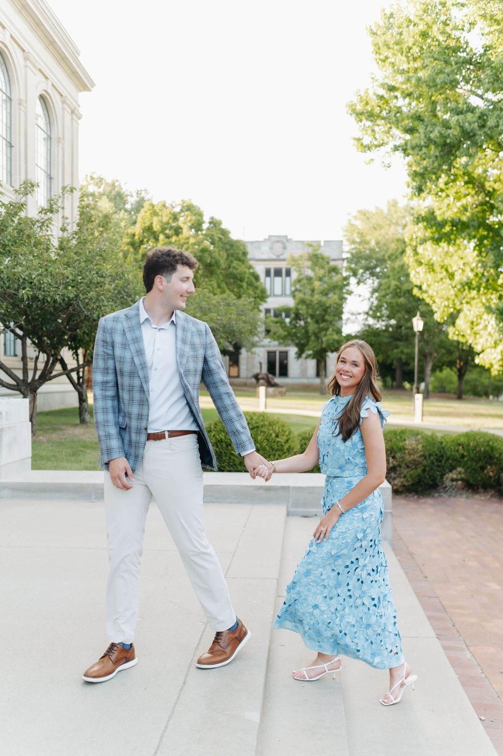 A couple holds hands and smiles while walking outdoors near a brick building, man in plaid blazer, woman in blue floral dress.