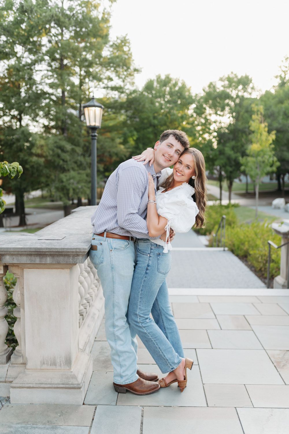 A couple embraces and smiles on a stone terrace surrounded by lush green trees during golden hour.