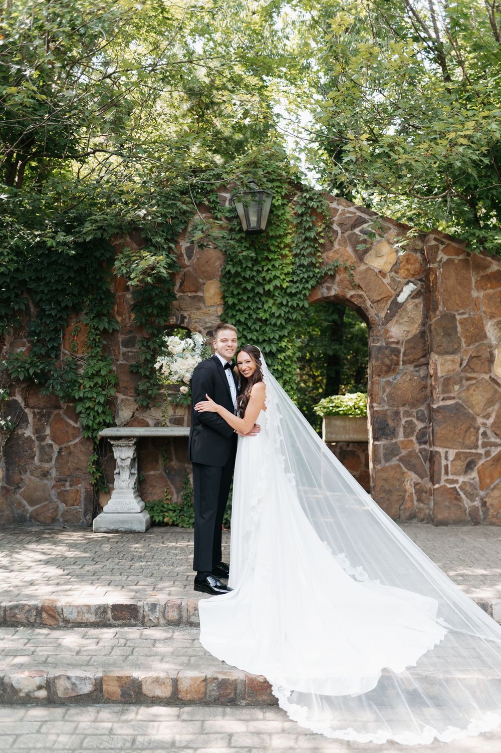 Bride and groom pose together in front of a stone arch with ivy, her cathedral veil flowing elegantly.