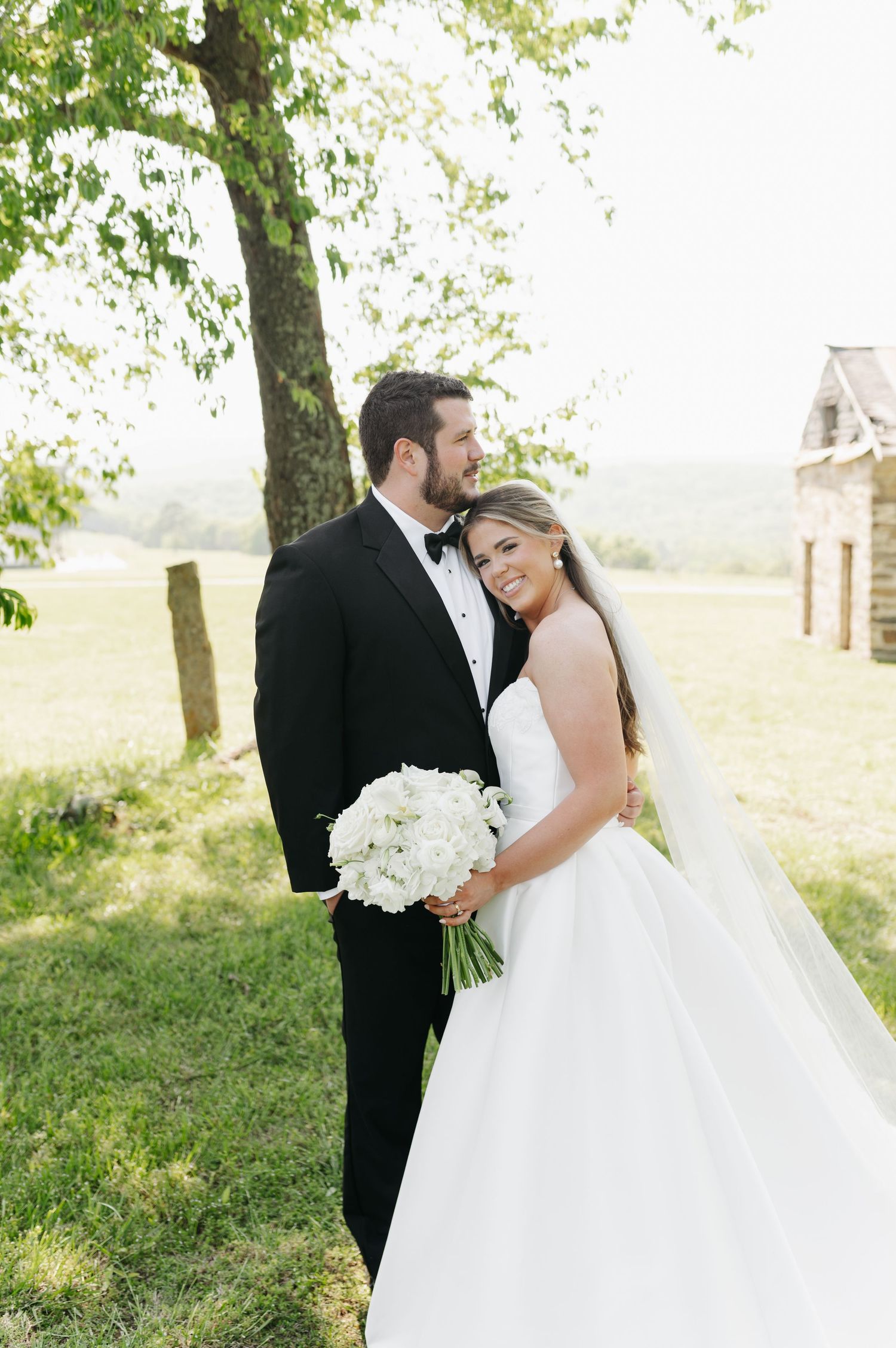 Bride and groom embracing outdoors under a tree, bride holding white floral bouquet in elegant wedding attire.