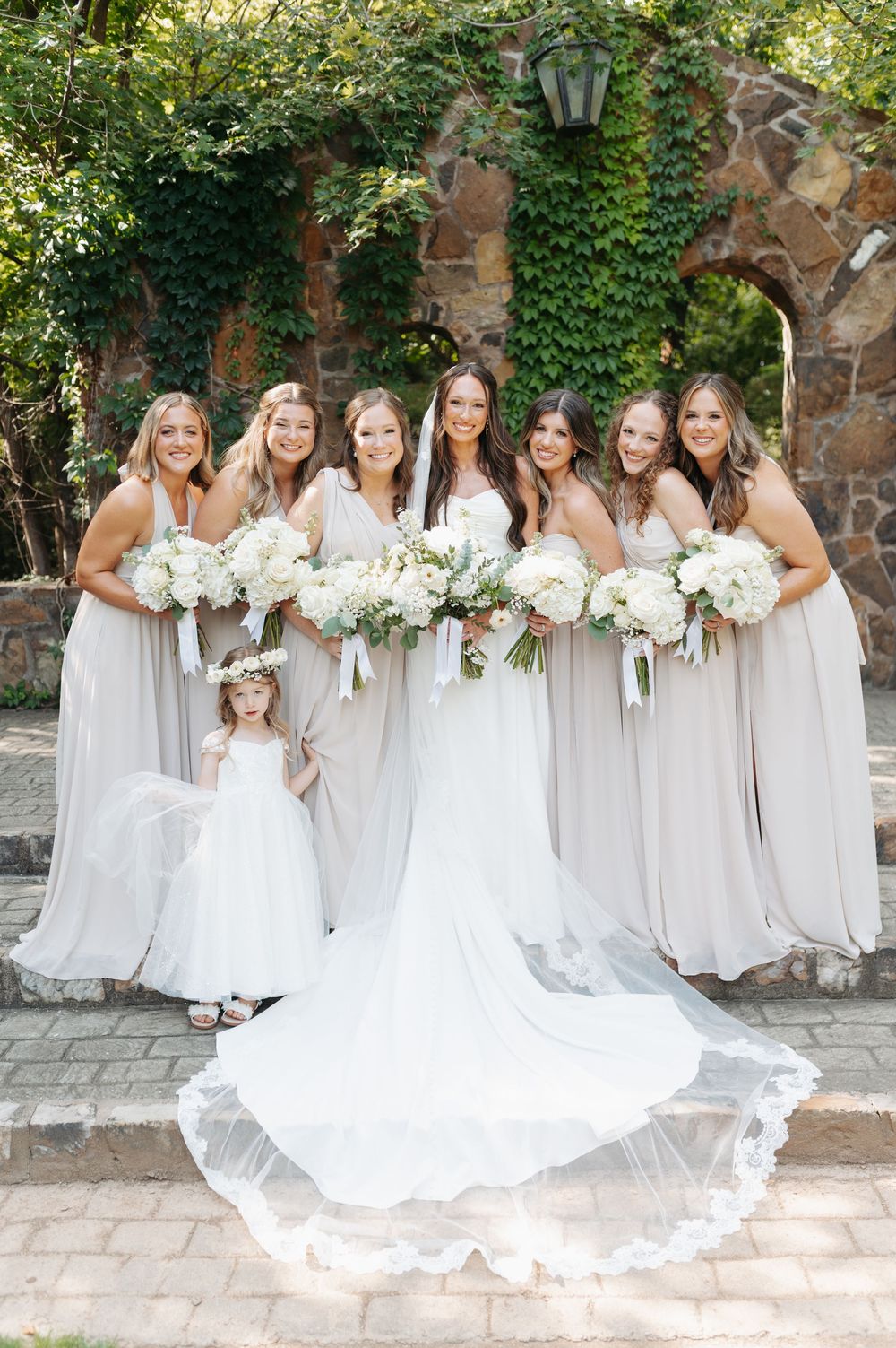 Bride in white gown with bridesmaids in taupe dresses holding white bouquets, posing with flower girl outdoors.