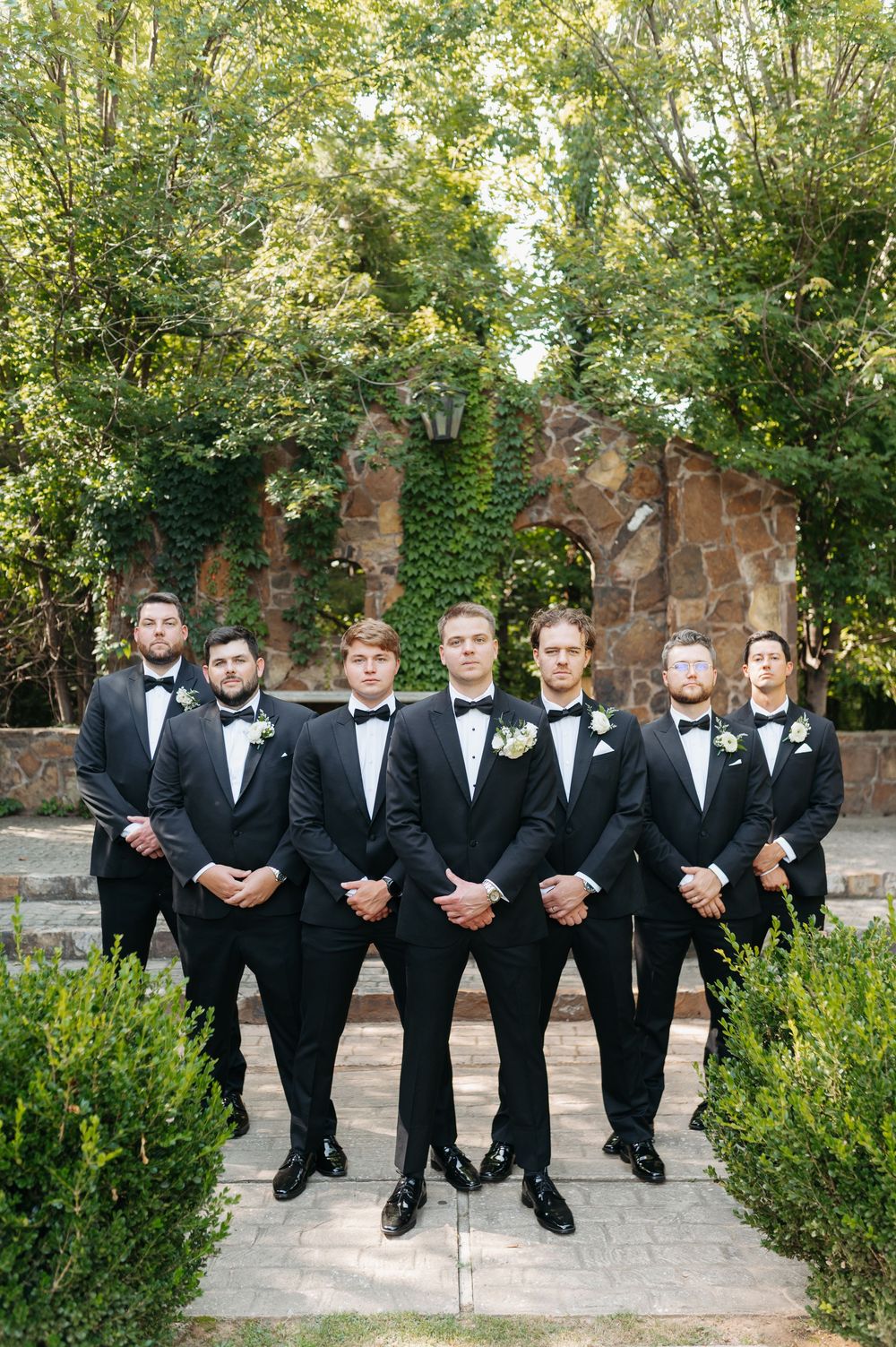 Six groomsmen in black tuxedos with bow ties and boutonnieres pose formally outdoors at a wedding venue.