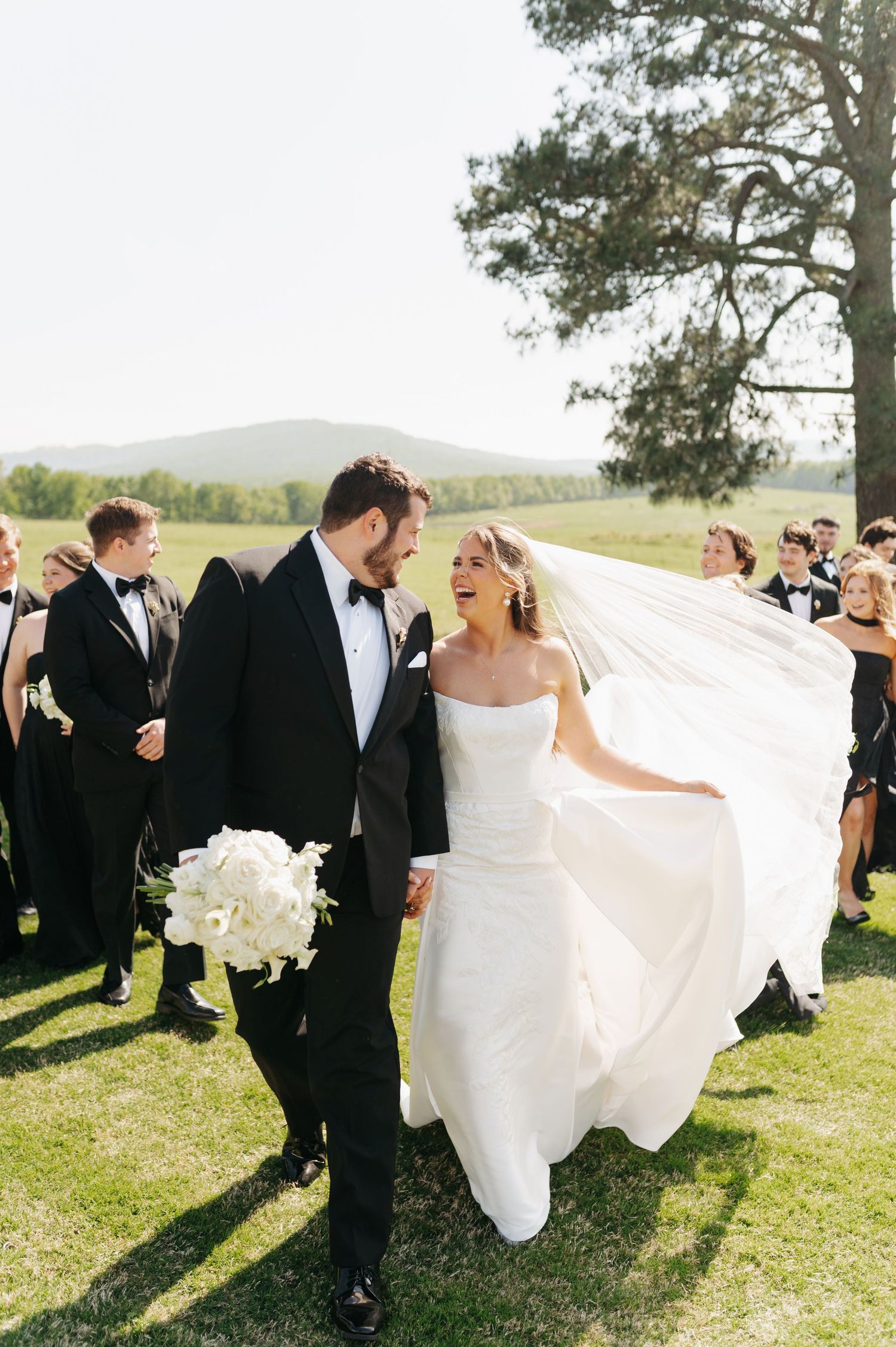 Bride and groom laugh together outdoors, her veil flowing in the wind, with wedding party in black tuxedos behind them.