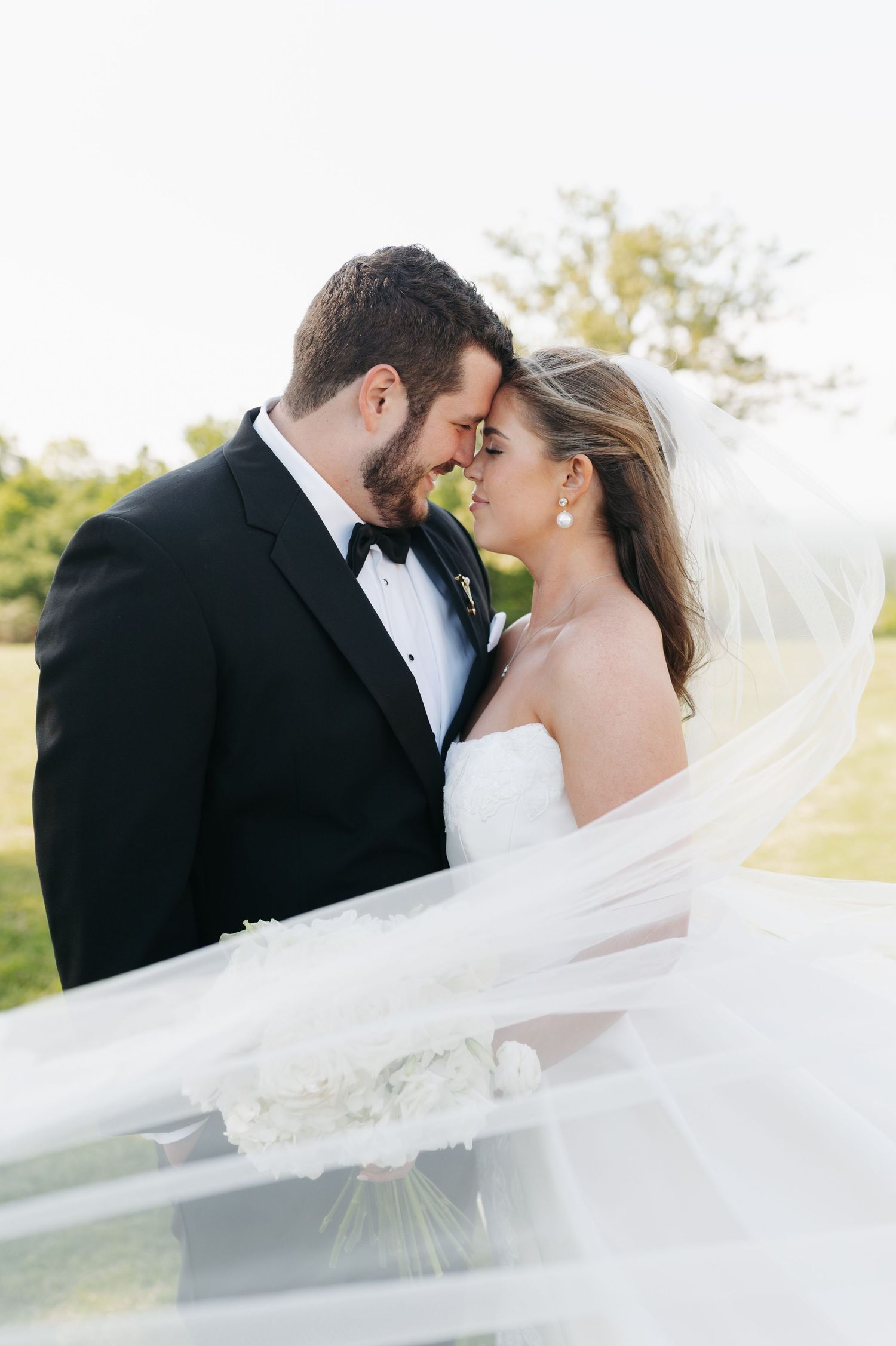 A bride and groom share a romantic forehead touch moment, her veil flowing gracefully in the breeze during outdoor wedding photos.