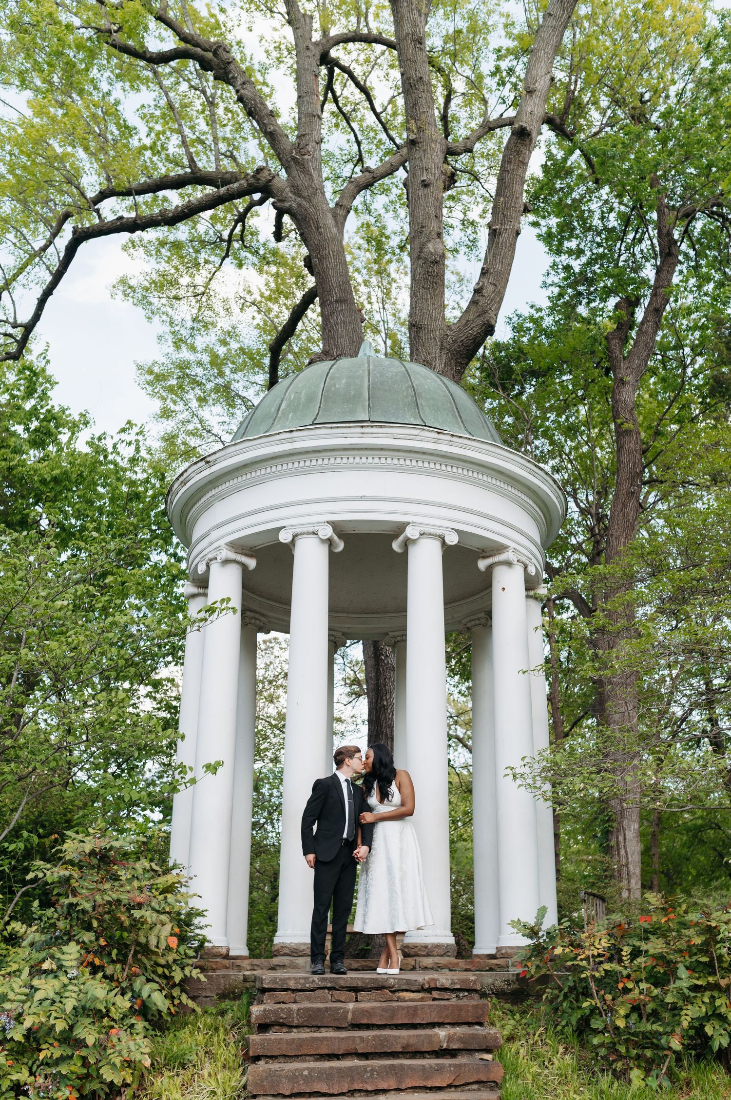 Couple sharing a romantic kiss under a white classical gazebo with columns surrounded by lush green trees.