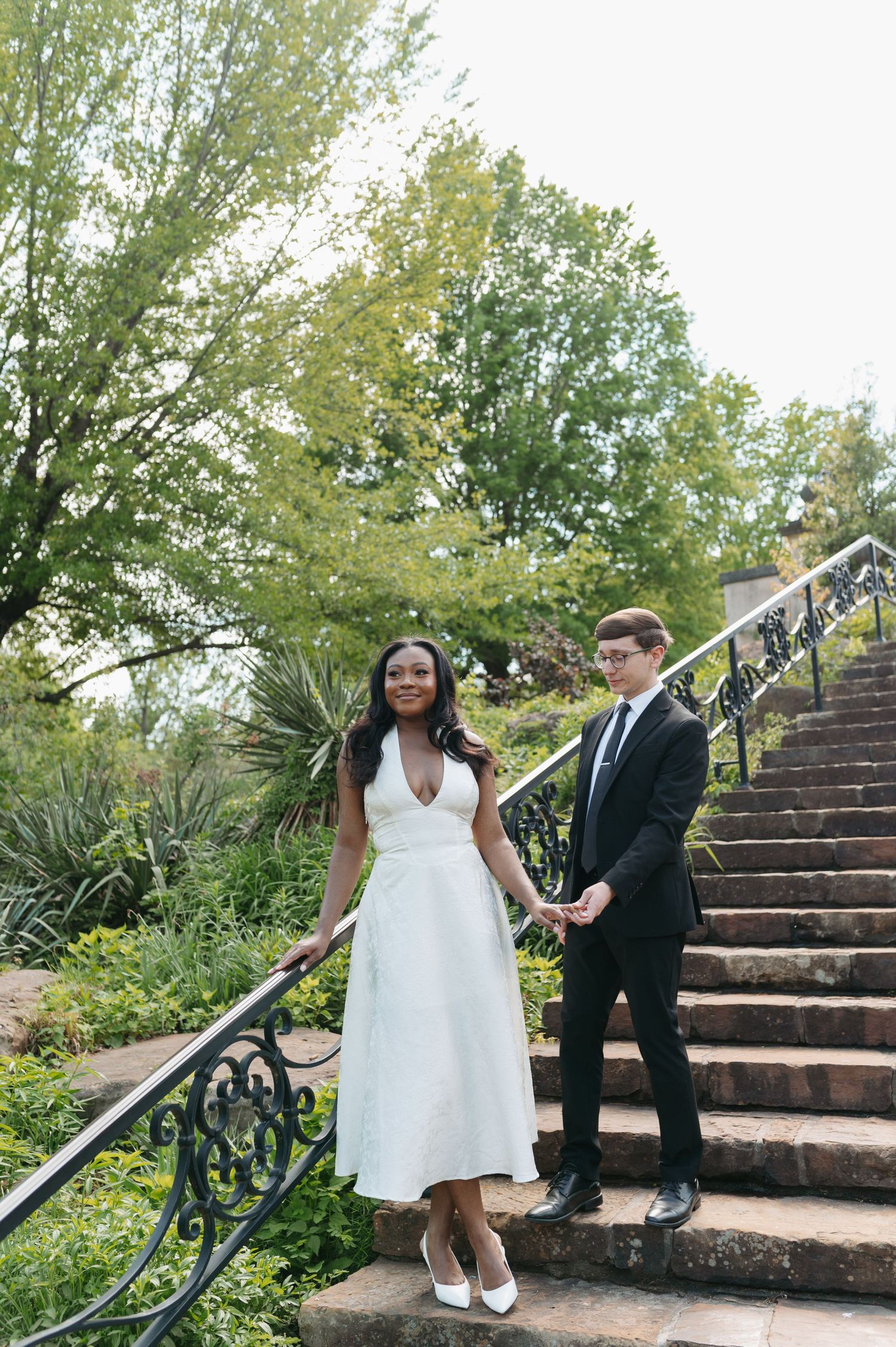 Interracial couple in wedding attire holding hands on stone steps surrounded by lush greenery.