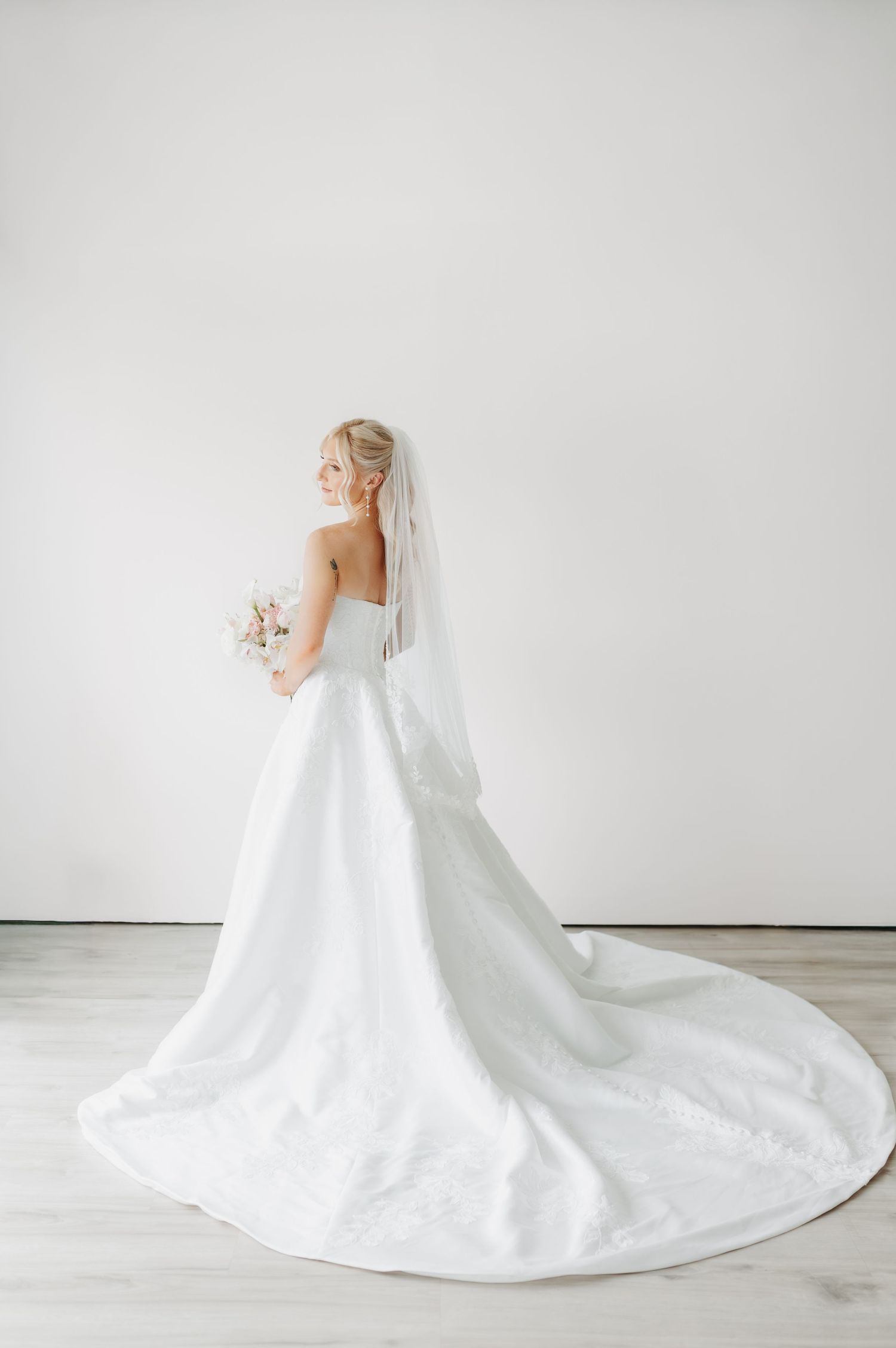 Bride in a strapless white ball gown with cathedral train and veil, holding a bouquet, posing with her back to camera.