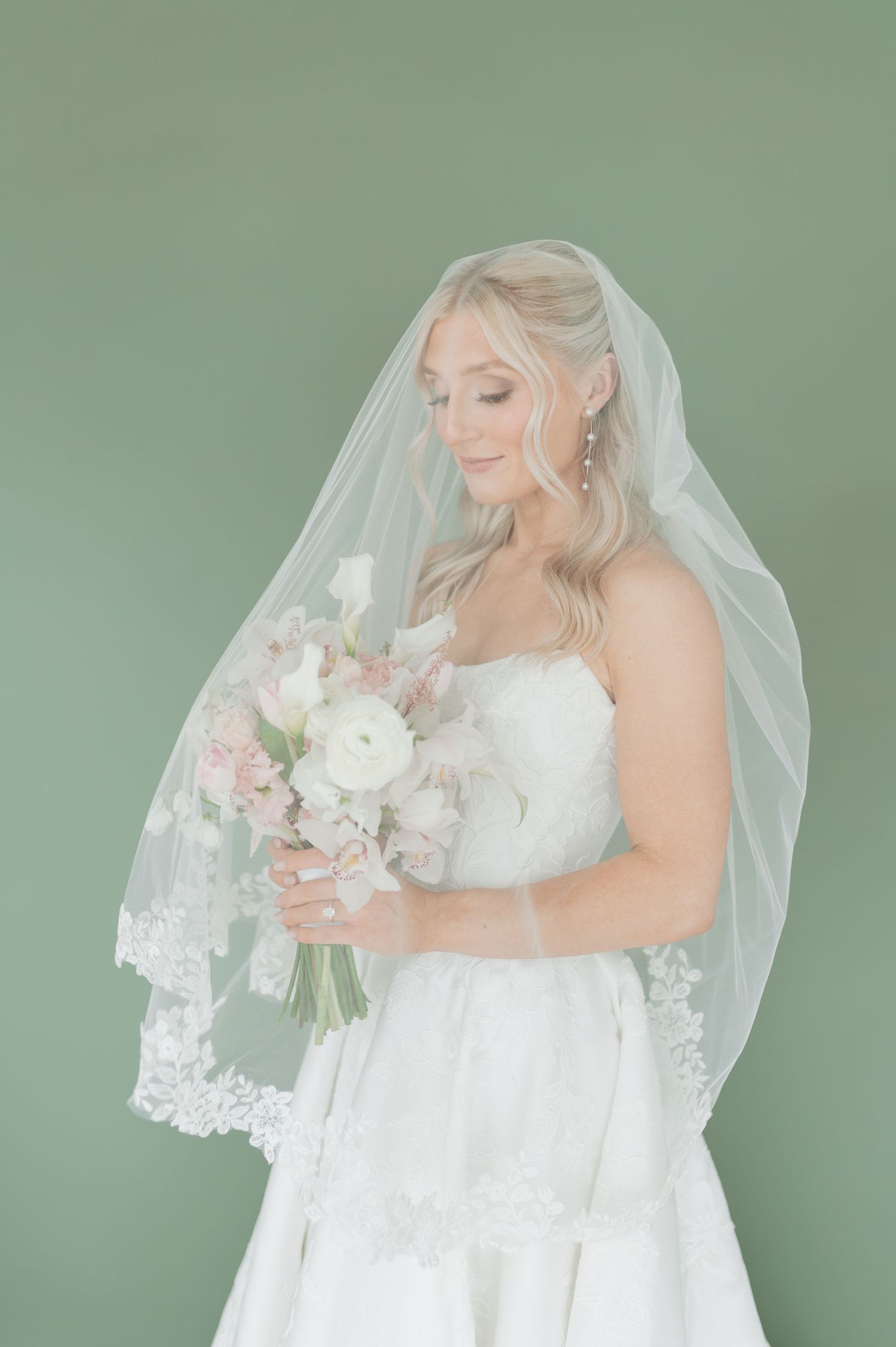 Bride in strapless white gown with lace-edged veil holding pink and white bouquet against sage green background.