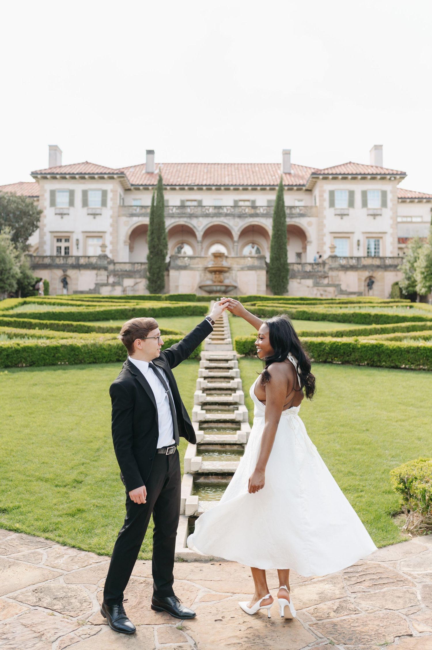 Bride and groom dancing together in formal attire before an elegant mansion with manicured gardens.