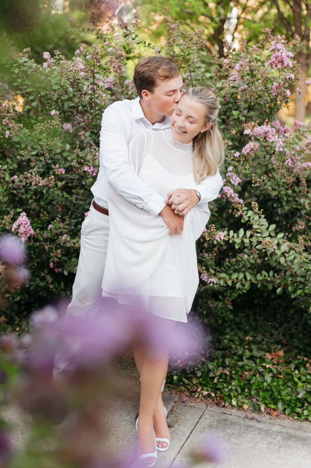 Couple in white outfits embracing among blooming pink flowers, man kissing woman on cheek in romantic garden setting.