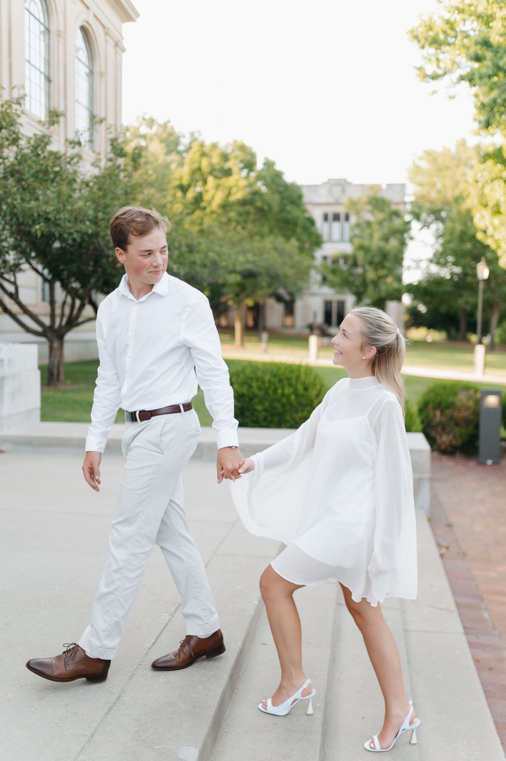 Couple in matching white outfits holding hands and walking on steps outdoors near a classic building.