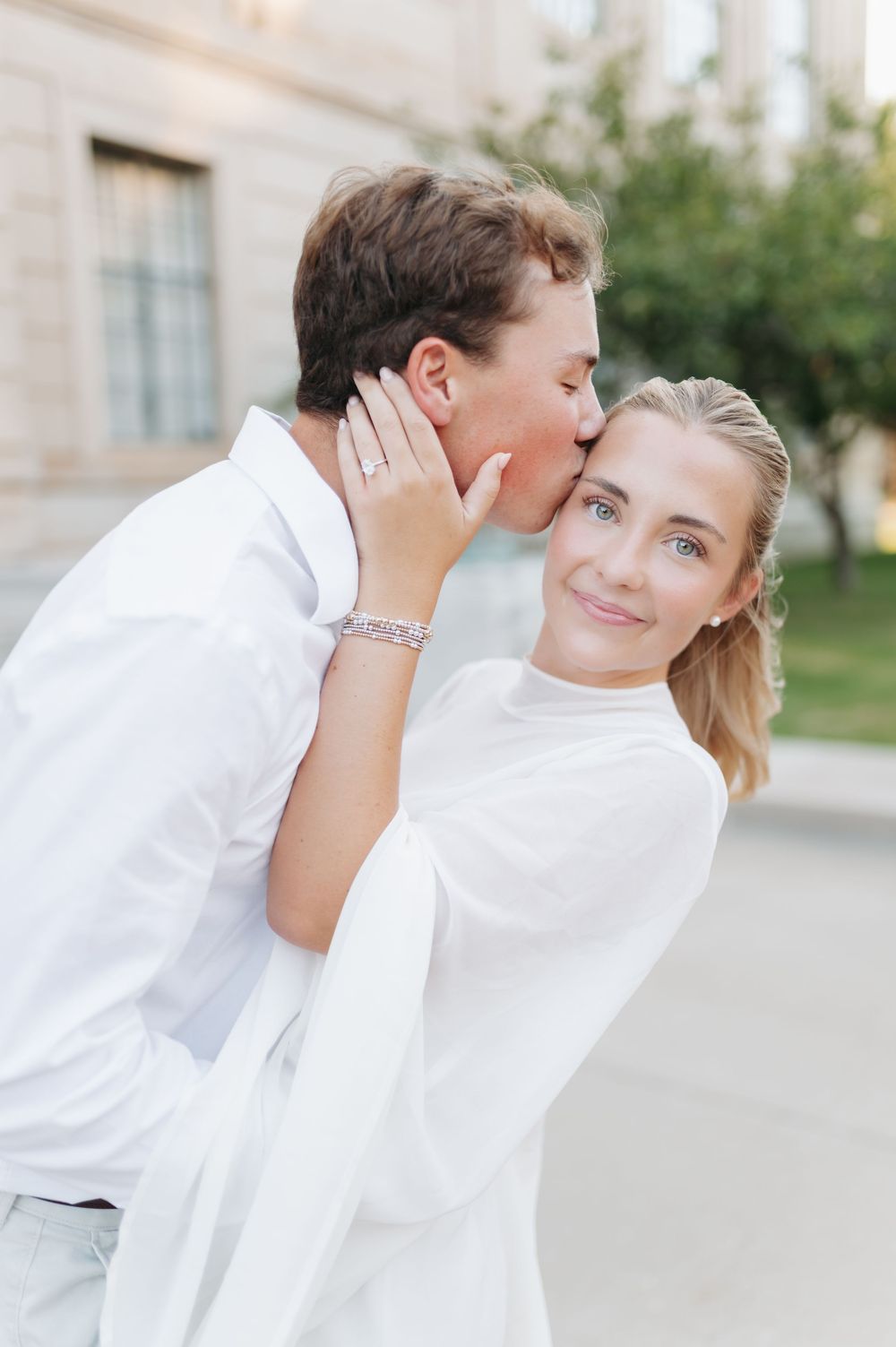 A man in white kisses a smiling blonde woman on the cheek outdoors during an engagement photo session.