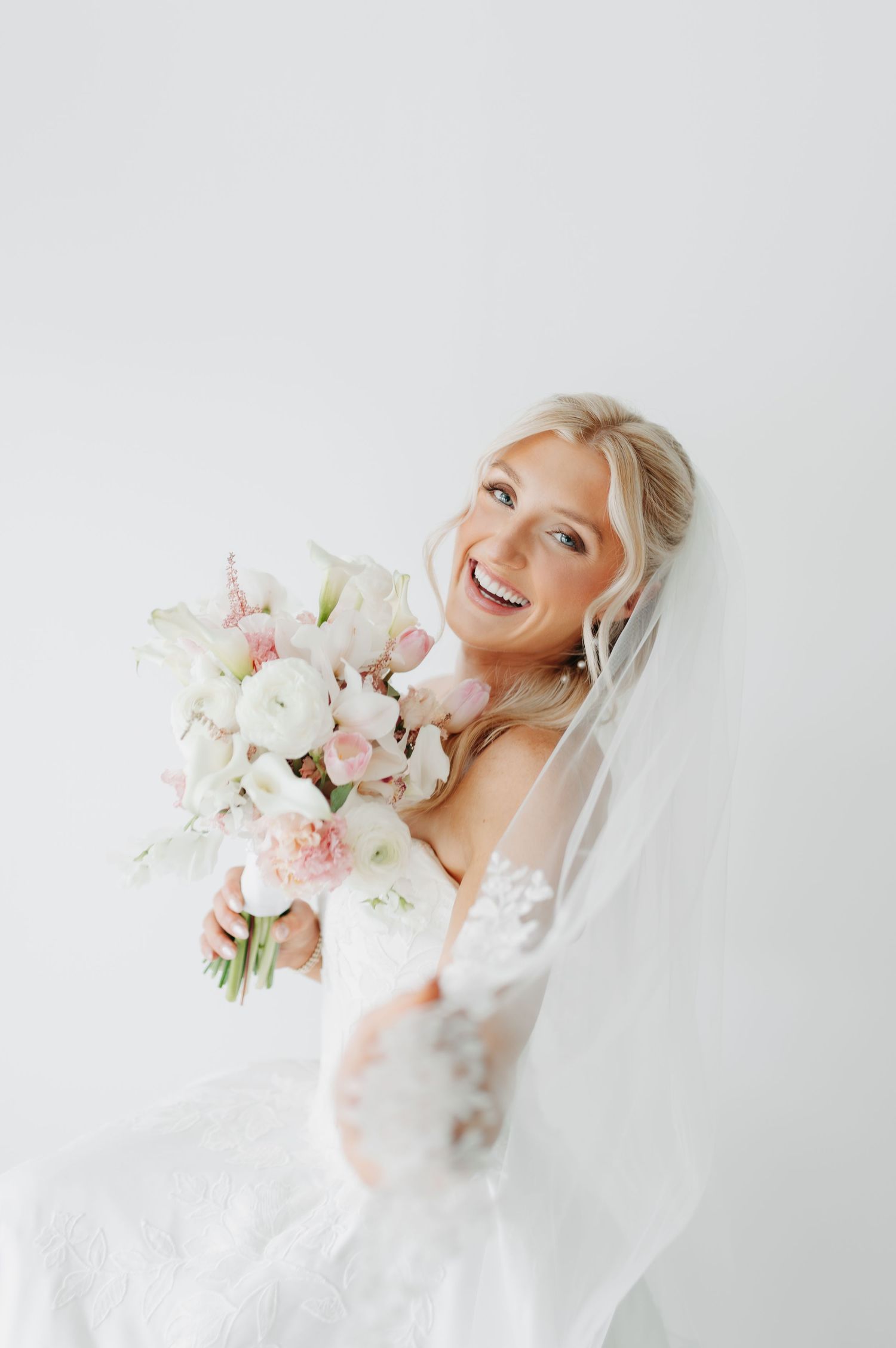 Smiling bride in lace gown holding pink and white floral bouquet with veil, against white background.