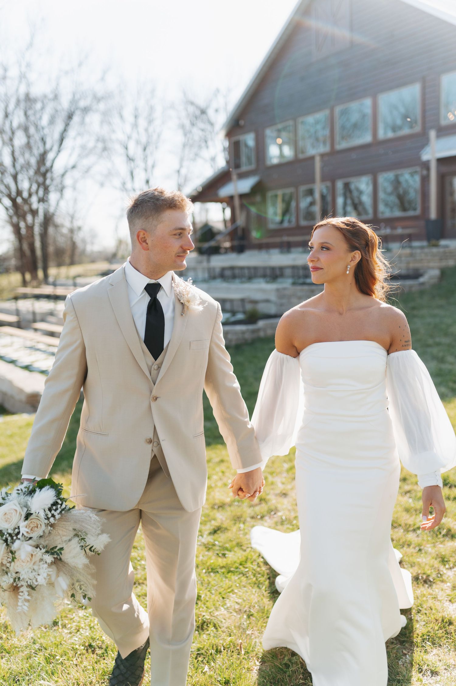 Bride in white off-shoulder gown and groom in tan suit hold hands walking outdoors near rustic wooden venue.