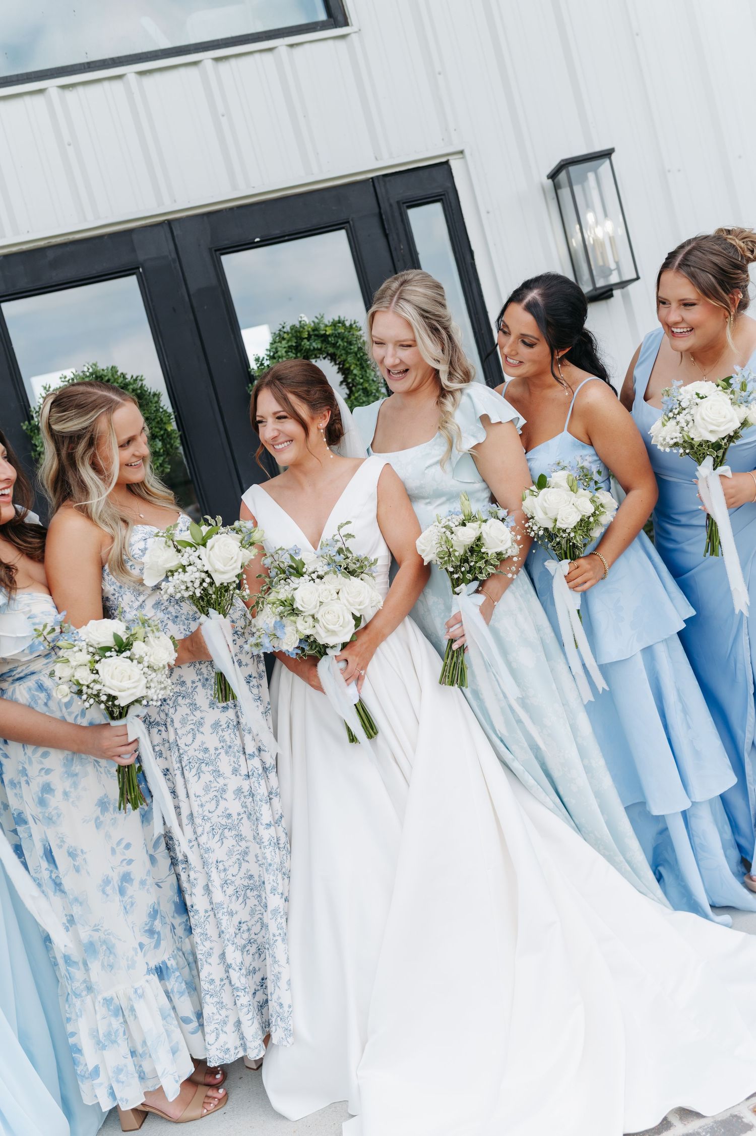Bride in white gown laughing with bridesmaids in blue dresses holding white bouquets outside a white venue.