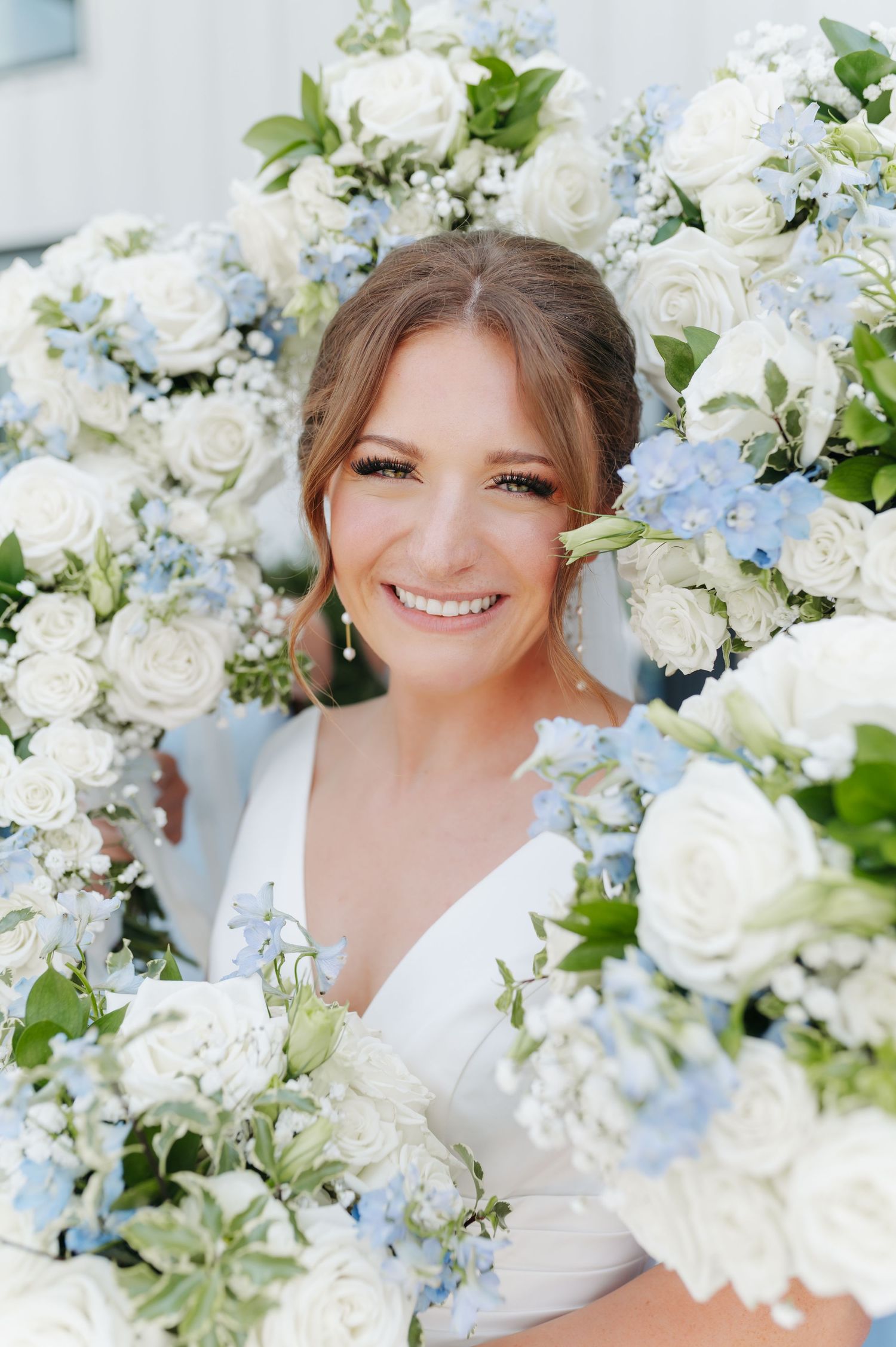 Smiling bride surrounded by lush white and blue floral bouquets featuring roses and delphinium on her wedding day.