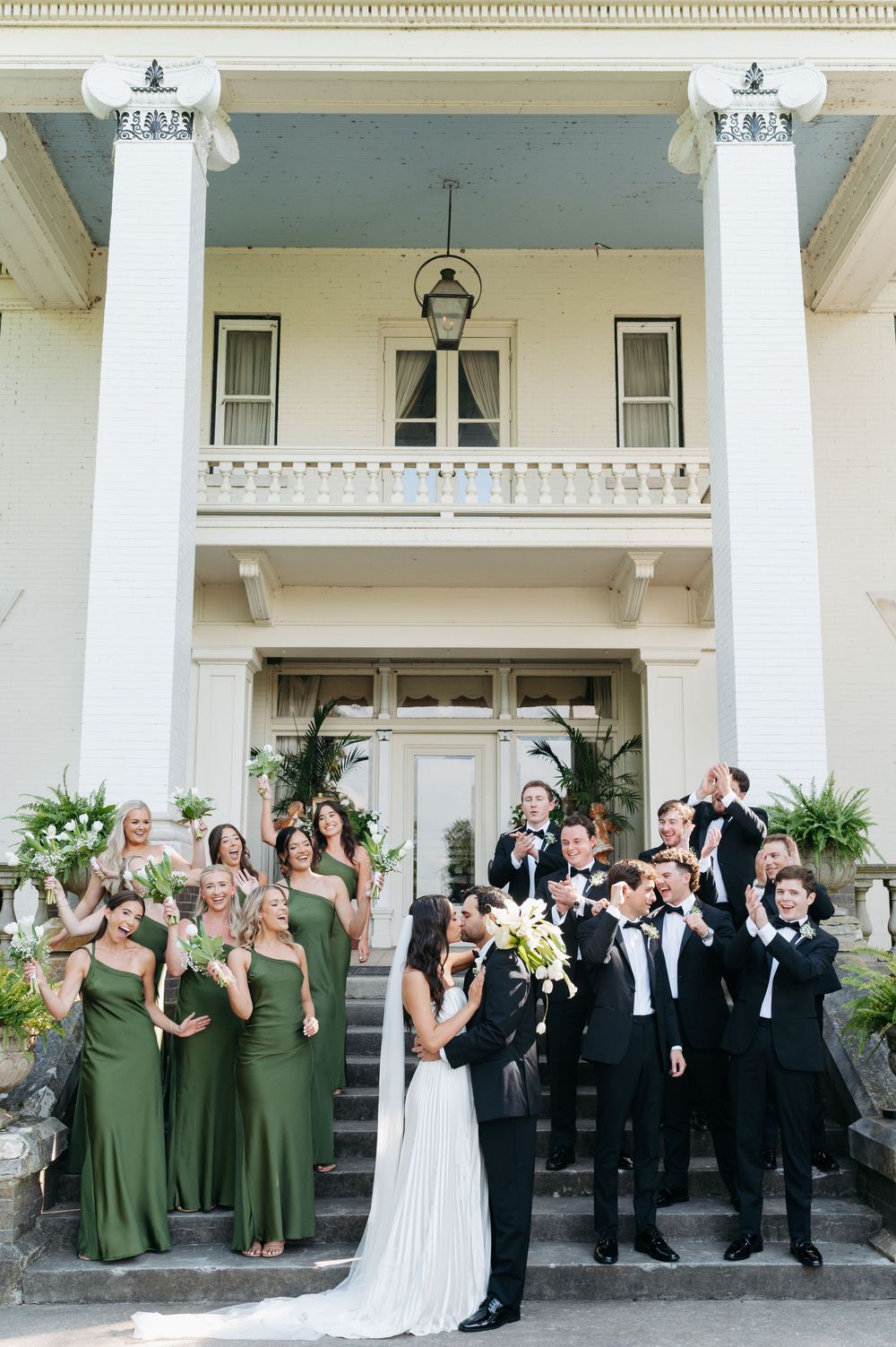 Bride and groom kiss on steps of white colonial mansion surrounded by wedding party in green gowns and black tuxedos.