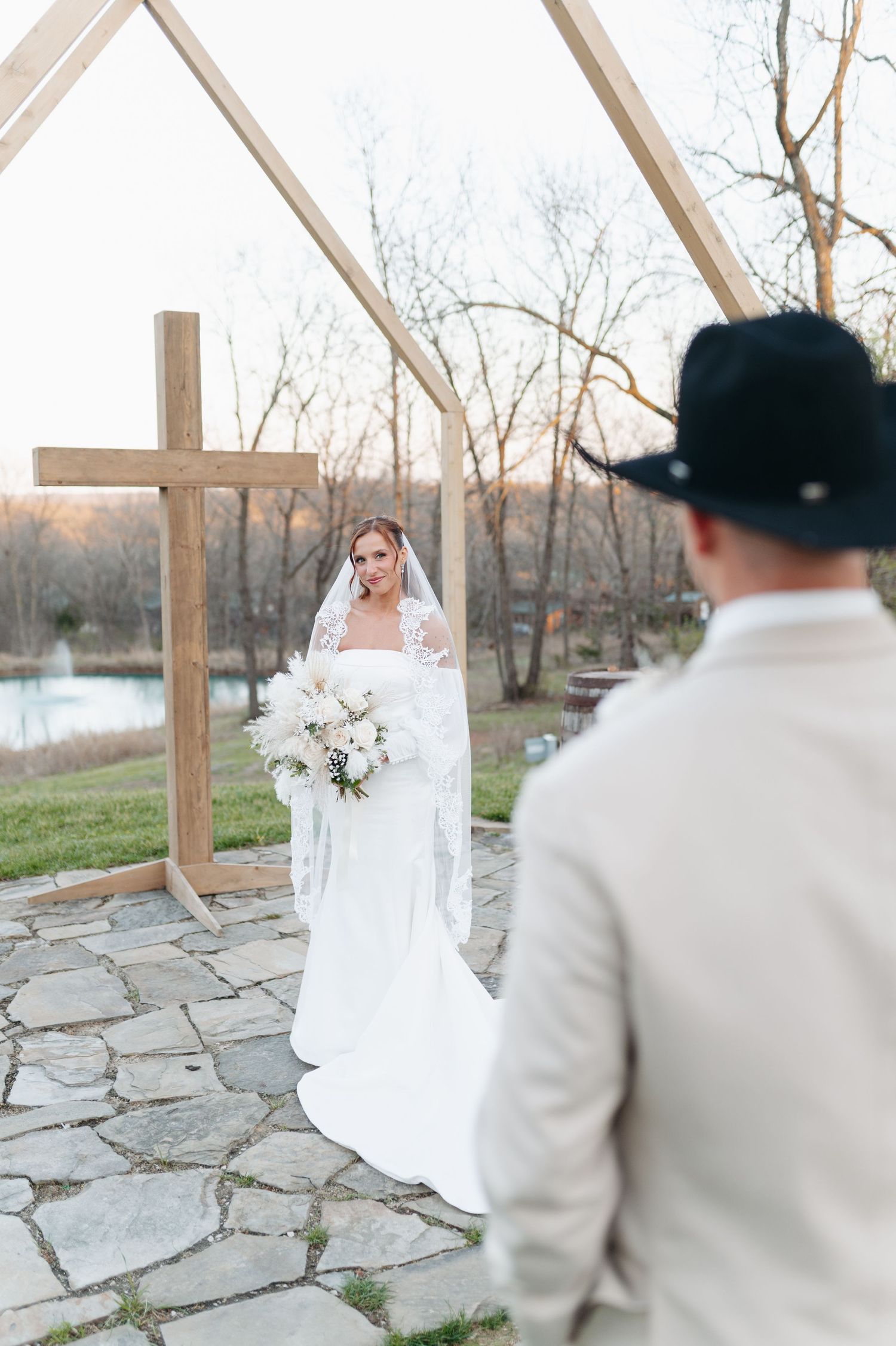 Bride in white gown and veil holds bouquet at outdoor wedding altar with wooden cross as groom in cowboy hat watches.