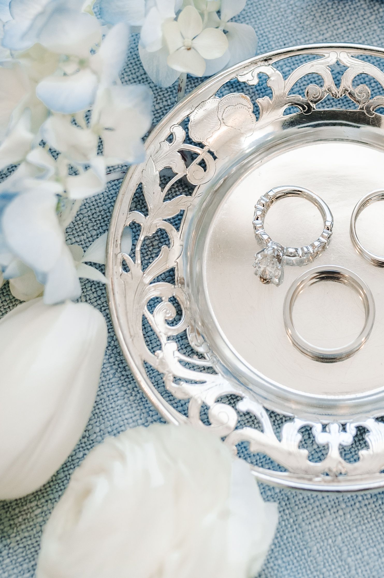 Silver filigree ring dish holding wedding bands and engagement ring, surrounded by white hydrangeas and tulips on blue linen.