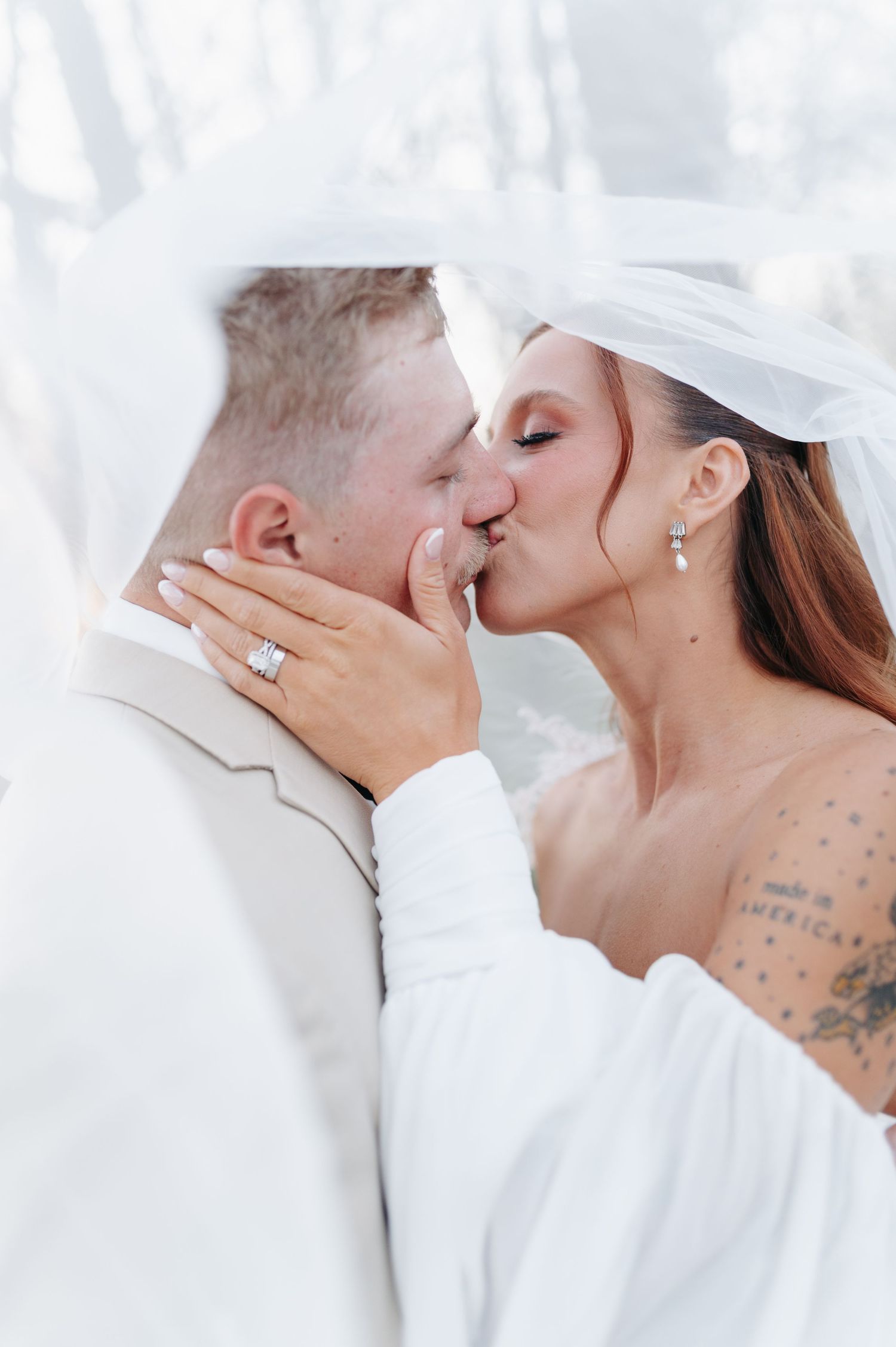 Bride and groom sharing a romantic kiss under a white veil, bride wearing a ring and off-shoulder dress.