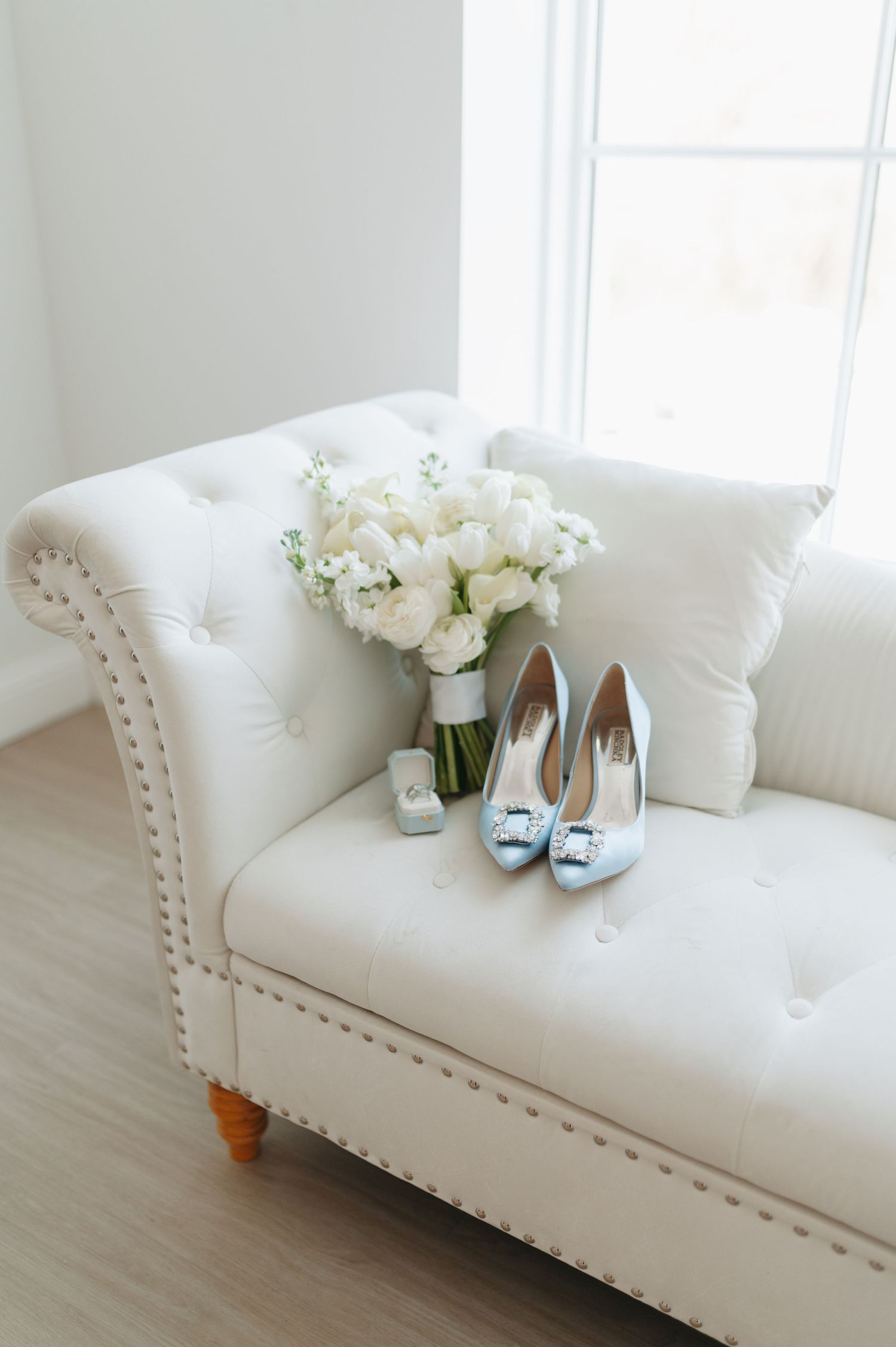 Elegant white tufted chaise lounge with light blue bridal heels, white floral bouquet, and ring box by a bright window.