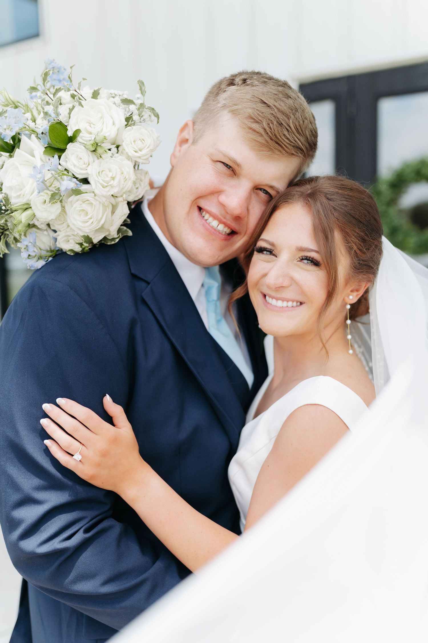 Smiling bride and groom embrace outdoors, bride holding white rose bouquet, groom in navy suit on wedding day.