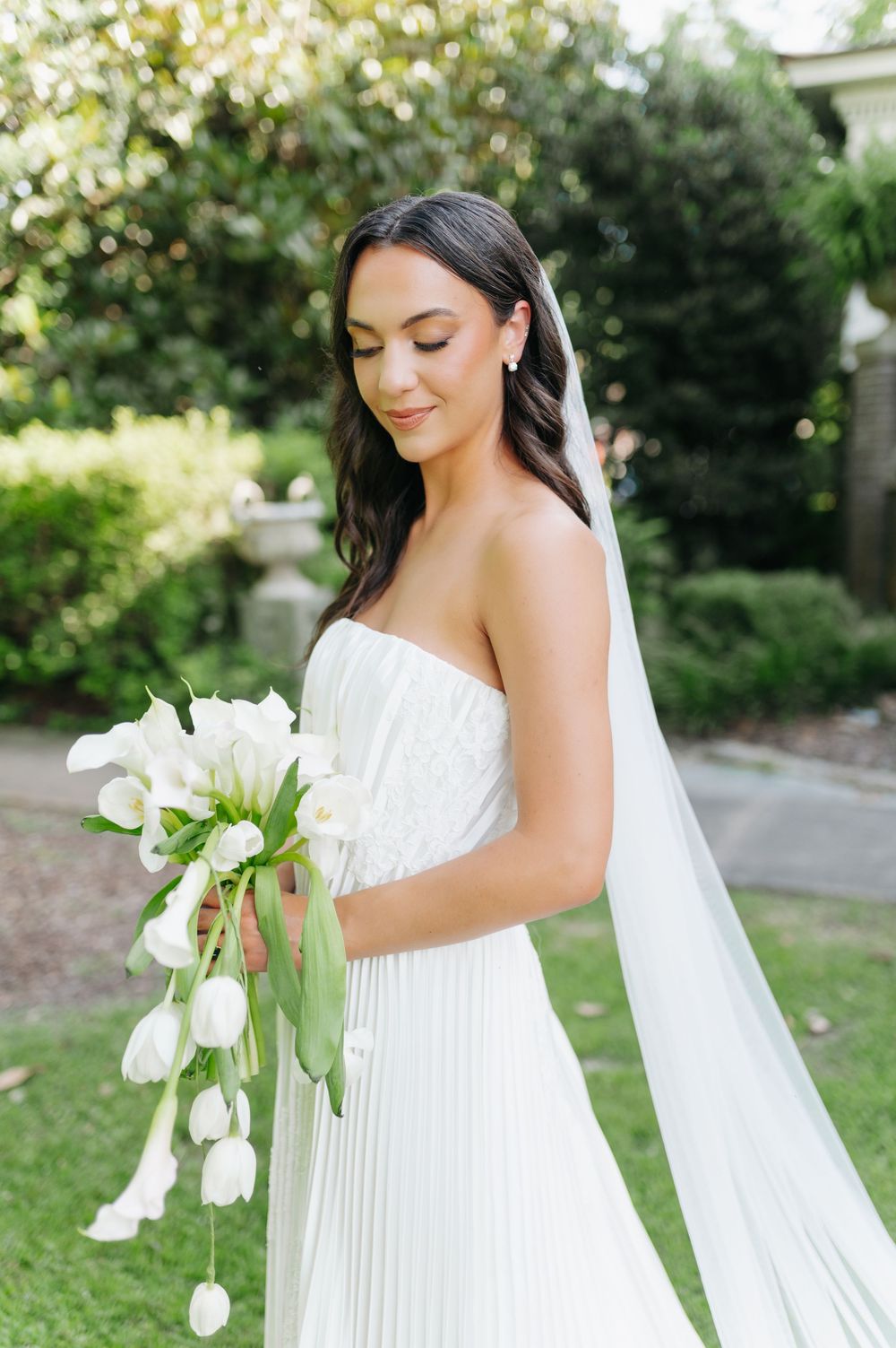 Bride in strapless pleated white gown holds cascading white tulip bouquet while wearing a veil in a lush garden.