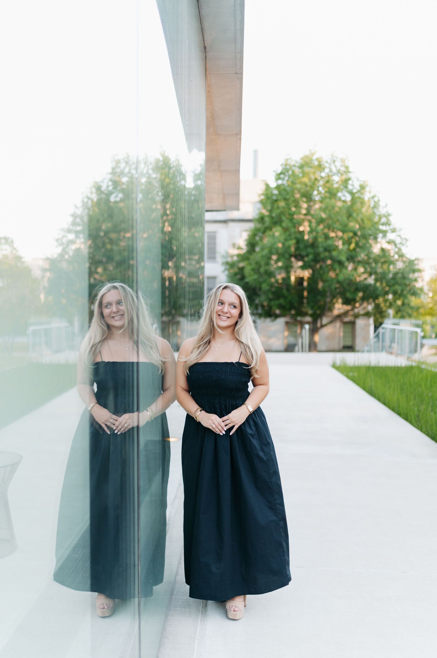 Young blonde woman in black maxi dress poses beside glass wall reflection outdoors near green trees.