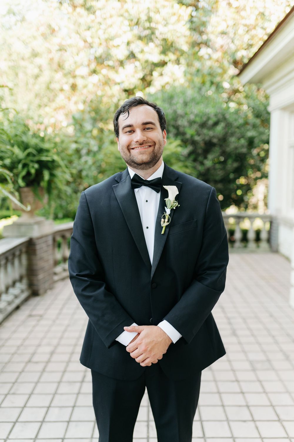 Smiling groom in black tuxedo with bow tie and white boutonniere stands on outdoor patio surrounded by lush greenery.