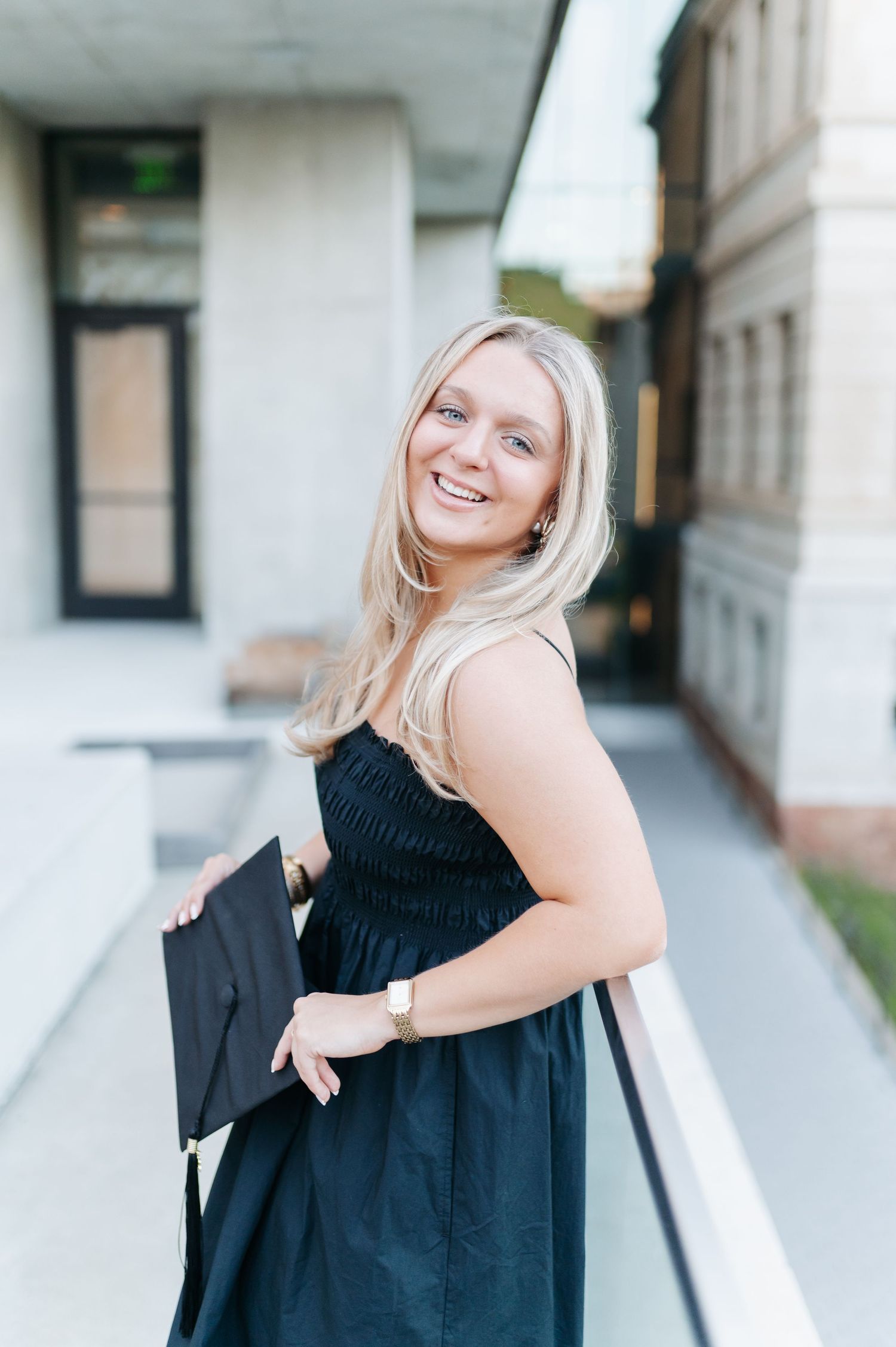 Smiling blonde woman in black dress holding graduation cap outside university building.