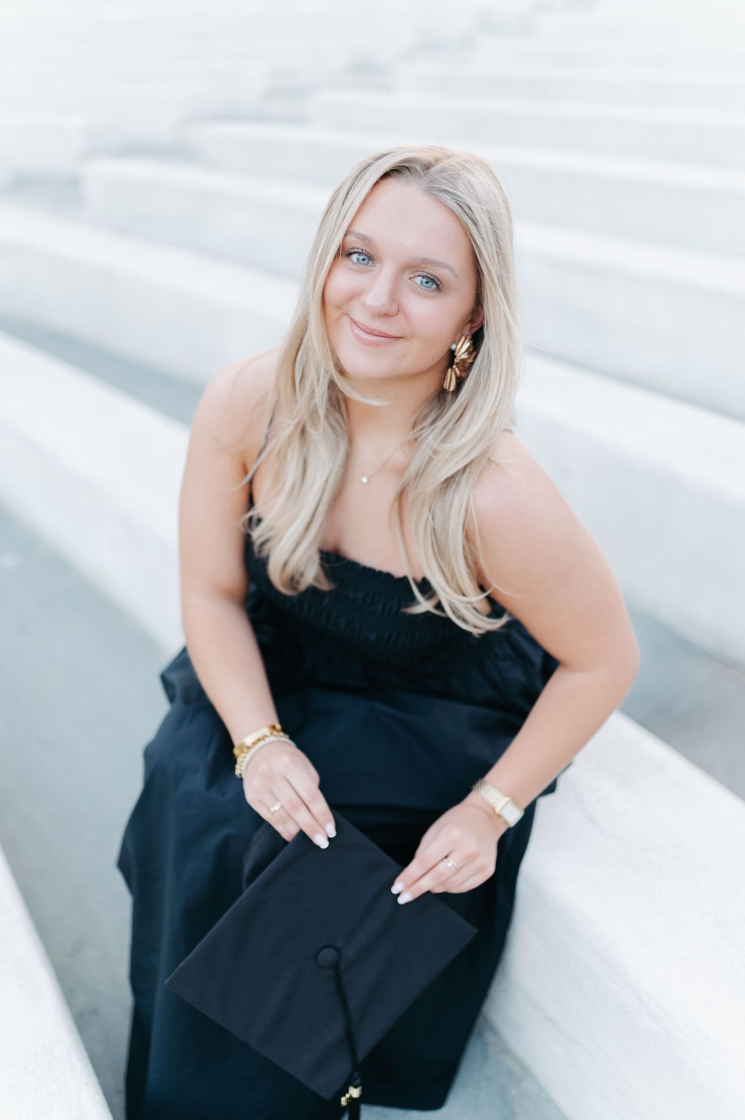 Smiling blonde woman in black strapless dress holding graduation cap while sitting on white marble steps.