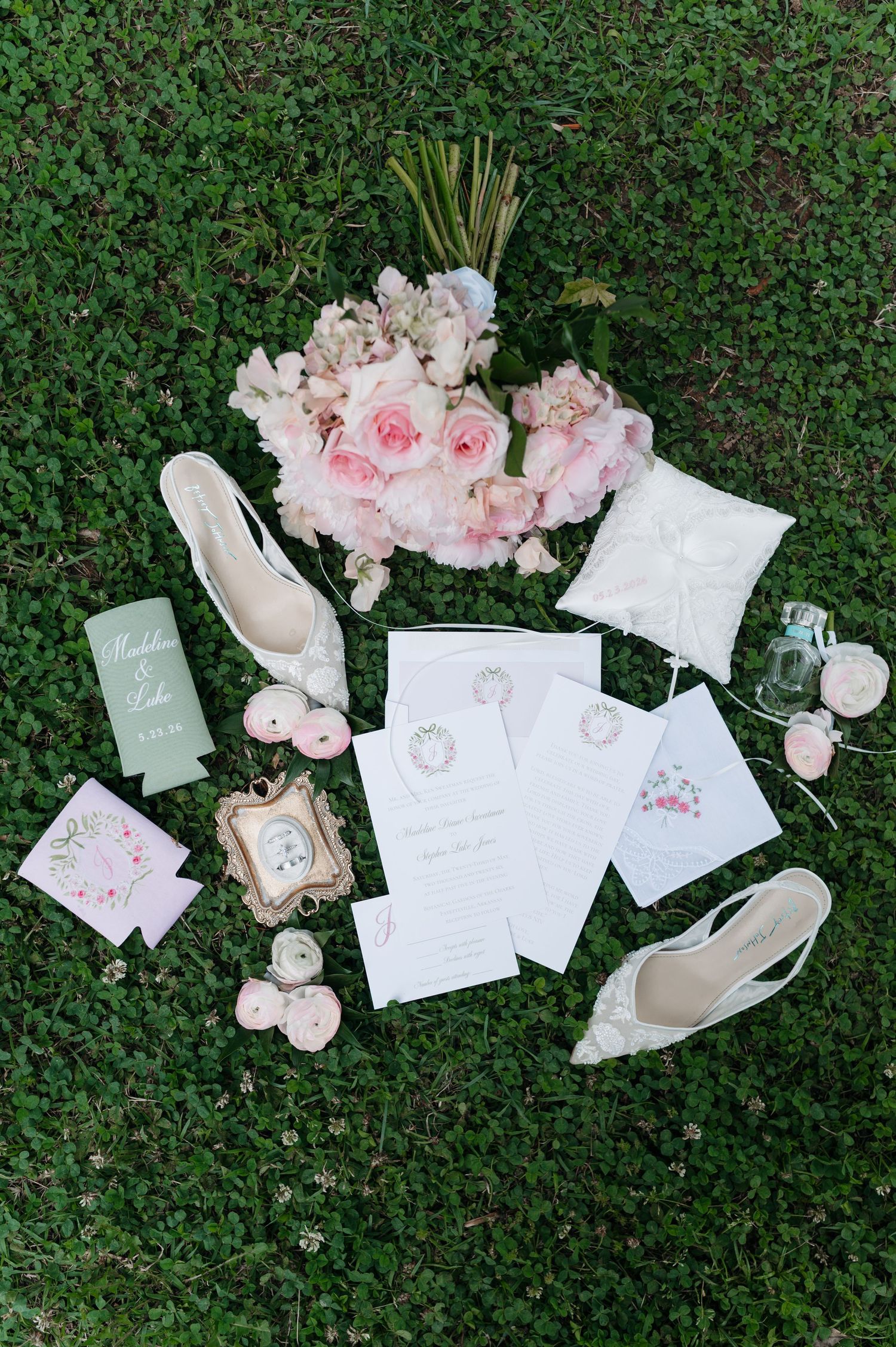 Elegant wedding flat lay with pink floral bouquet, lace heels, invitation suite, ring pillow, and accessories on green grass.