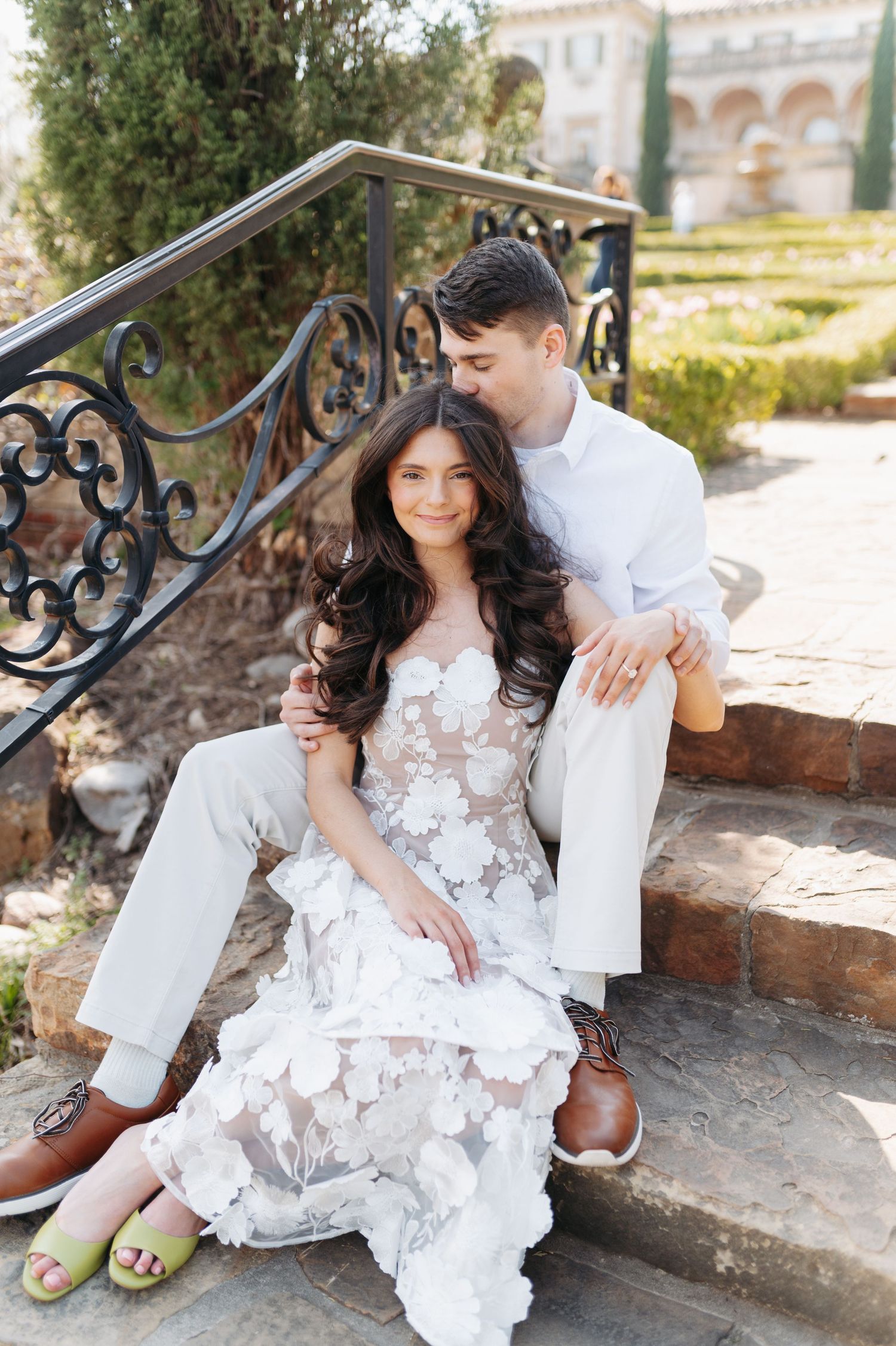Couple in white wedding attire sitting on stone steps, woman in floral lace gown, man in white suit kissing her head.