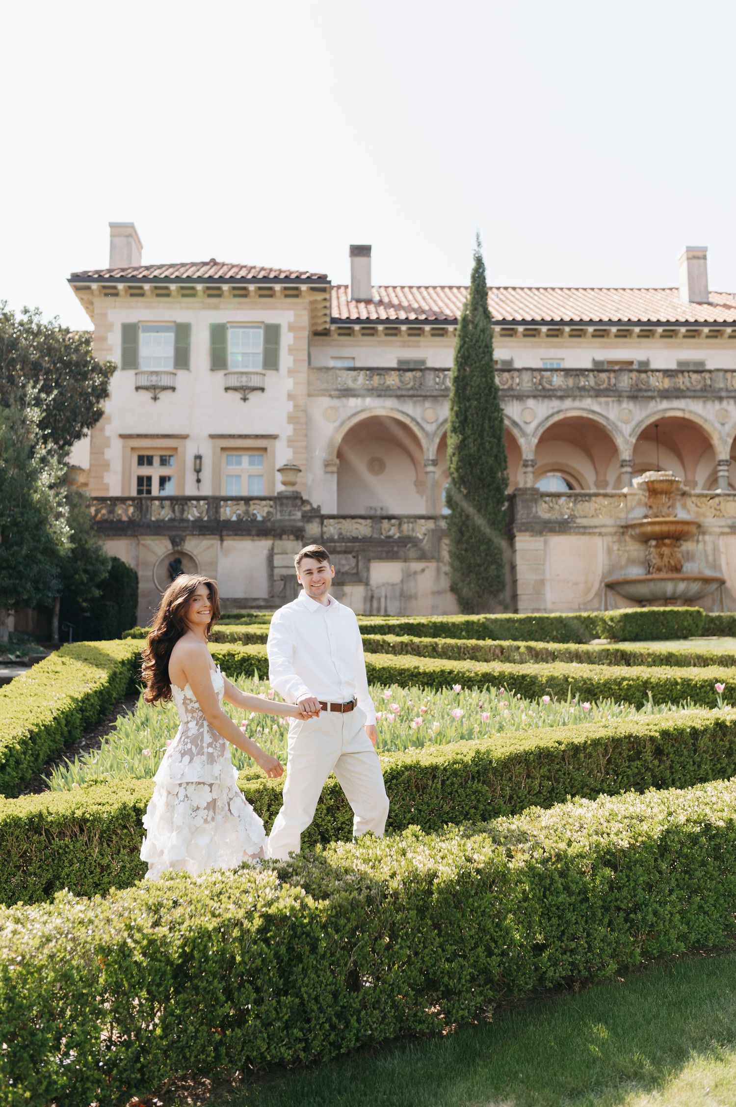 Couple in white wedding attire walks hand-in-hand through elegant formal garden with Italian villa backdrop.
