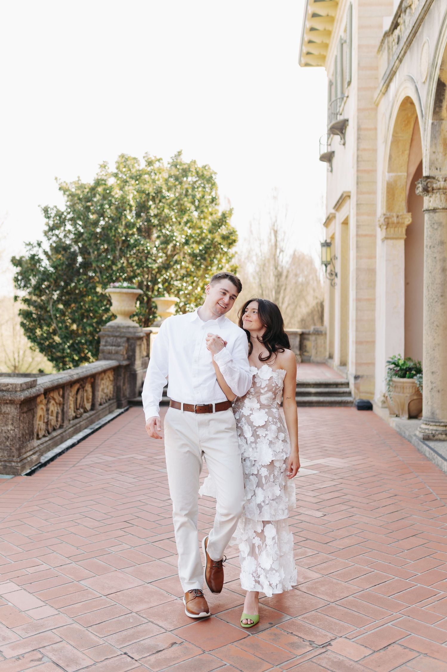 Couple walking together on elegant terrace, woman in floral dress leaning on man in white suit.