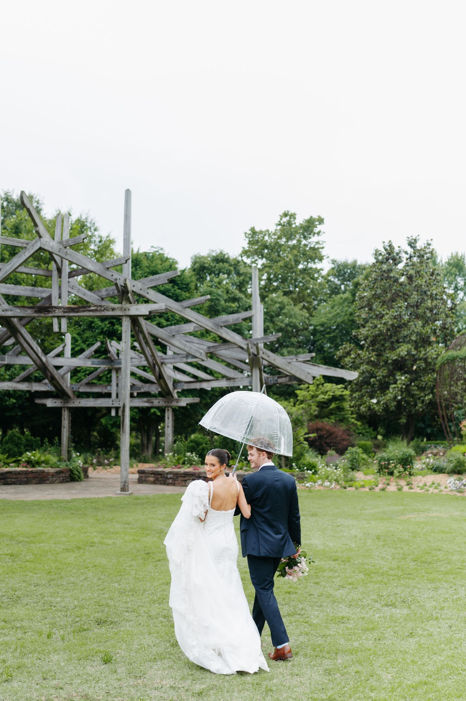 Bride and groom walking under a clear umbrella on a lush green lawn with a wooden pergola structure behind them.