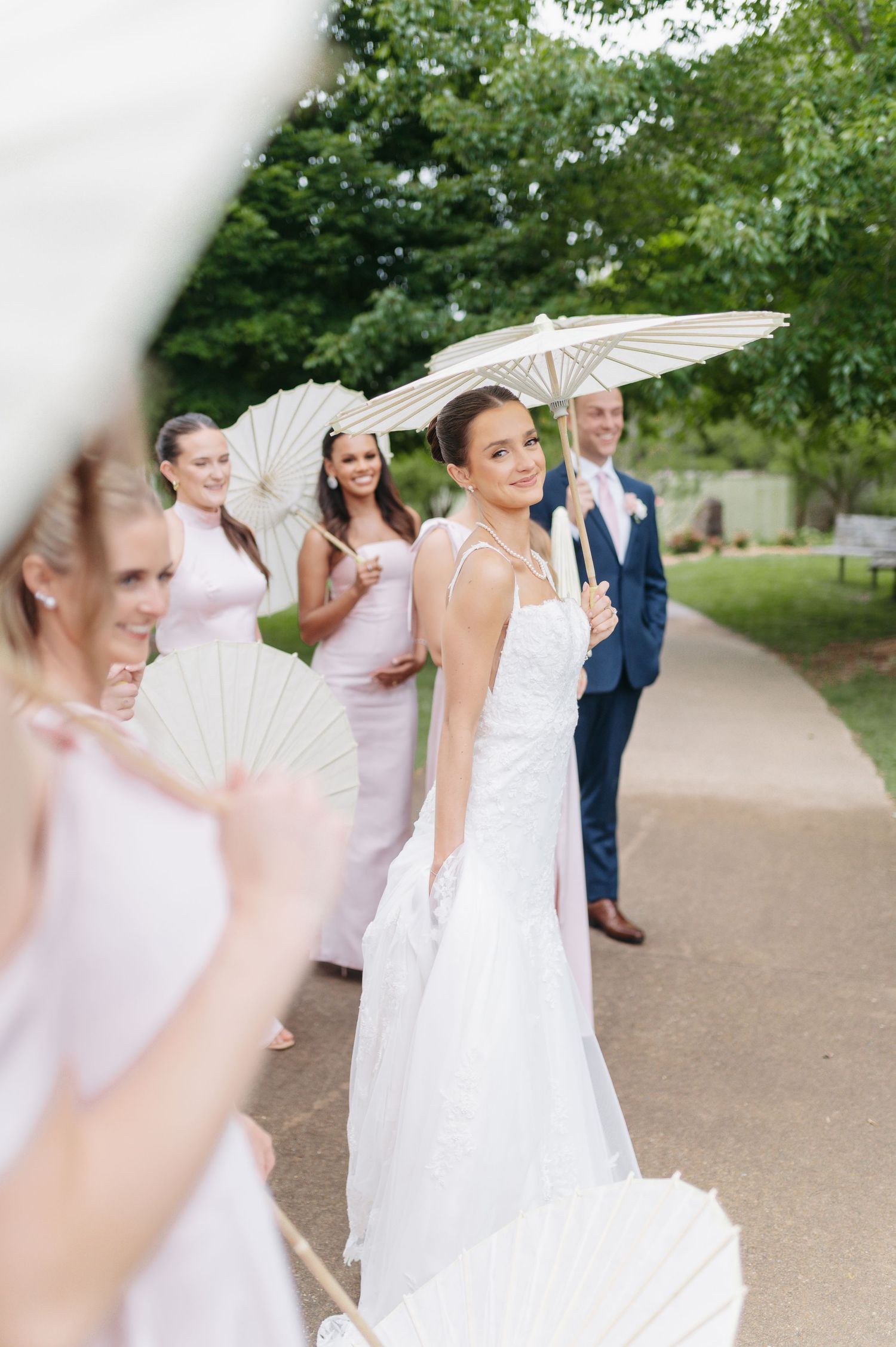 Bride in white gown holds parasol with bridesmaids in pink dresses and groomsman in blue suit outdoors.