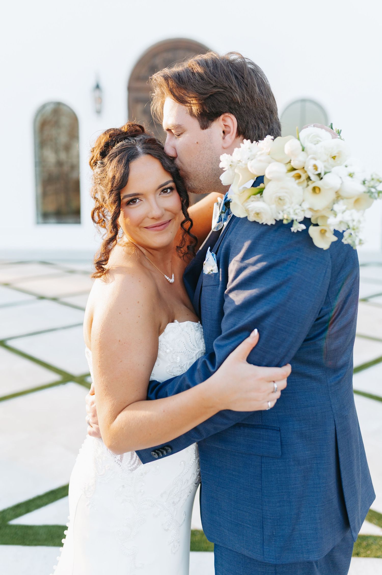 Bride in strapless lace gown smiles as groom in navy suit kisses her cheek, holding white floral bouquet outdoors.