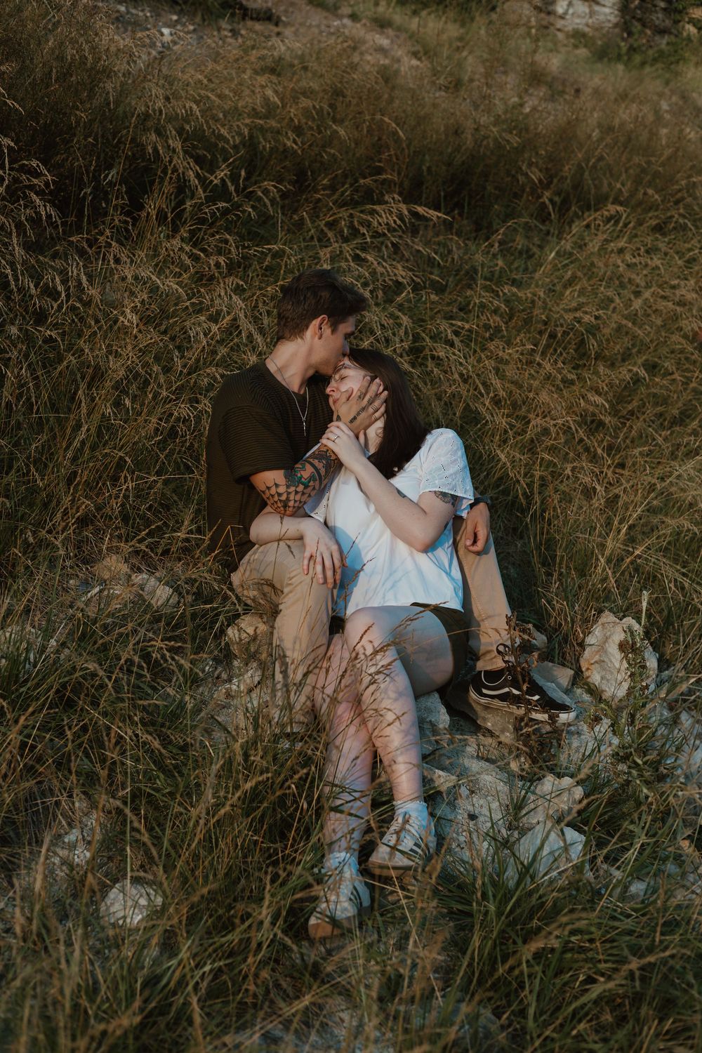 A couple relaxes together in tall grass during a romantic outdoor photoshoot at golden hour.