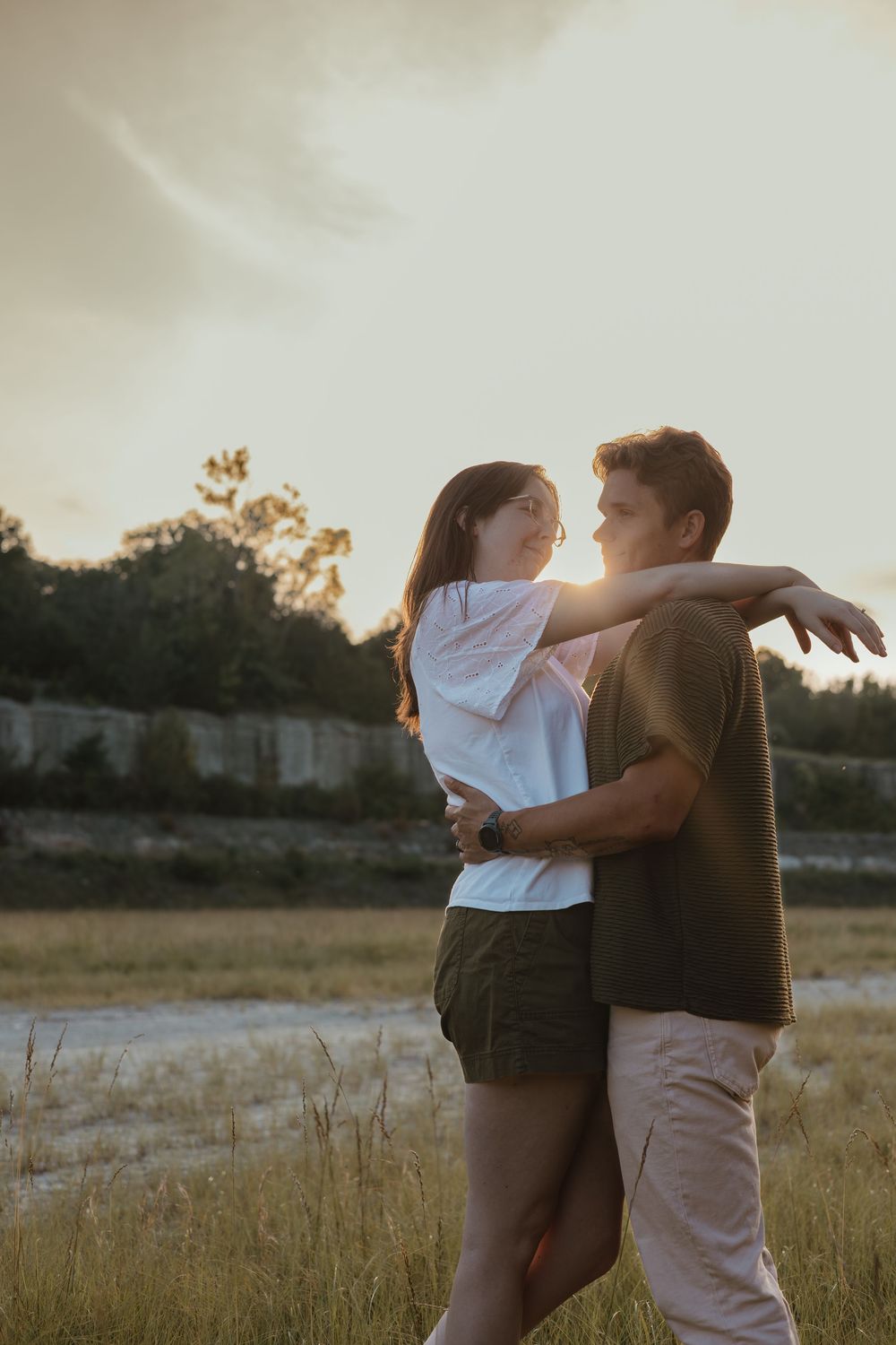 A couple embraces in a sunlit field during golden hour as sunlight streams through their silhouettes.