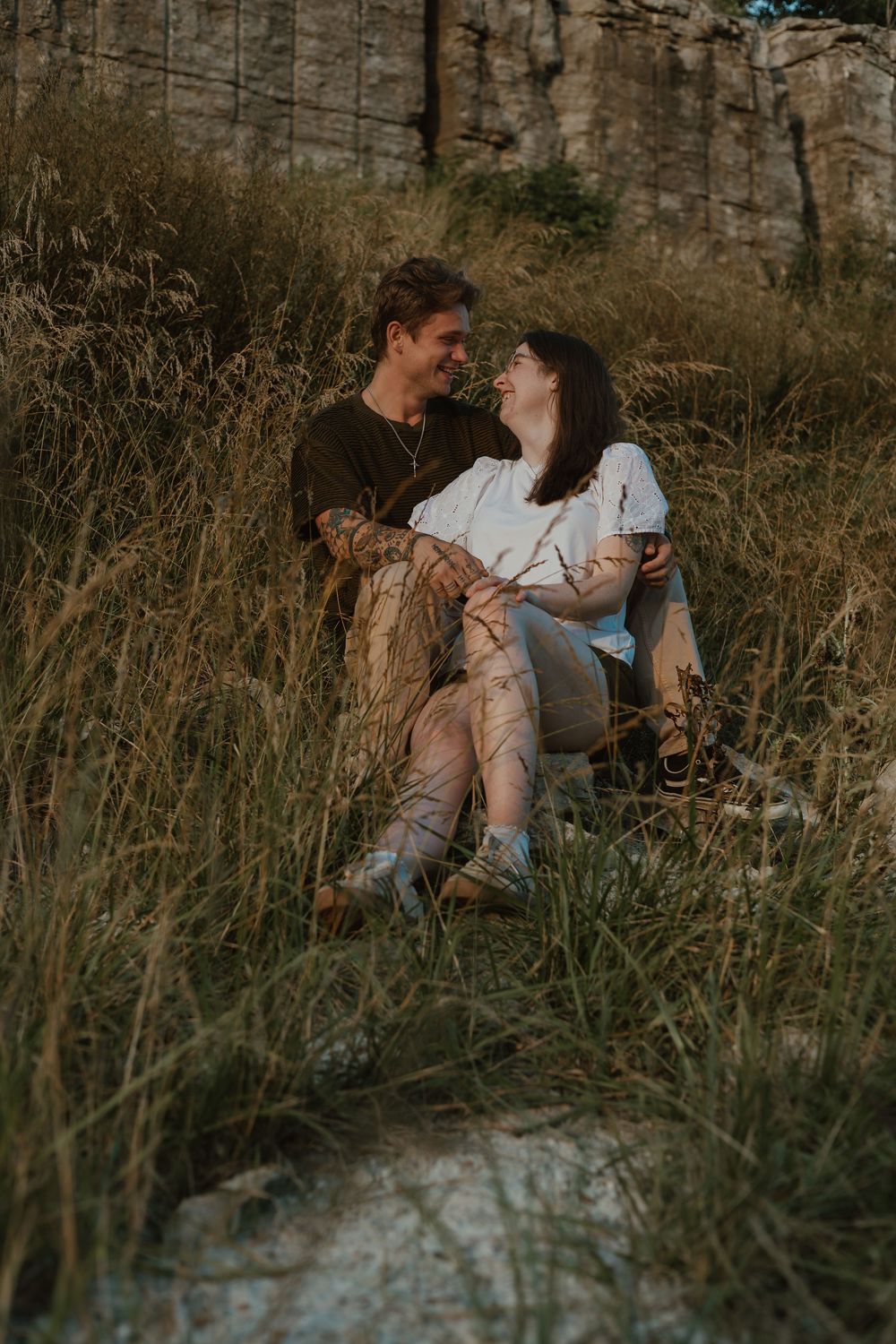 A romantic couple embraces while lying in tall grass during a moody outdoor photoshoot at sunset.