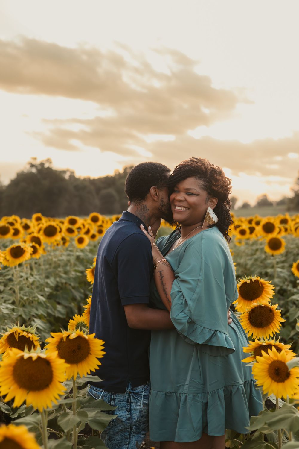 A couple stands lovingly embraced in a golden sunflower field at sunset.