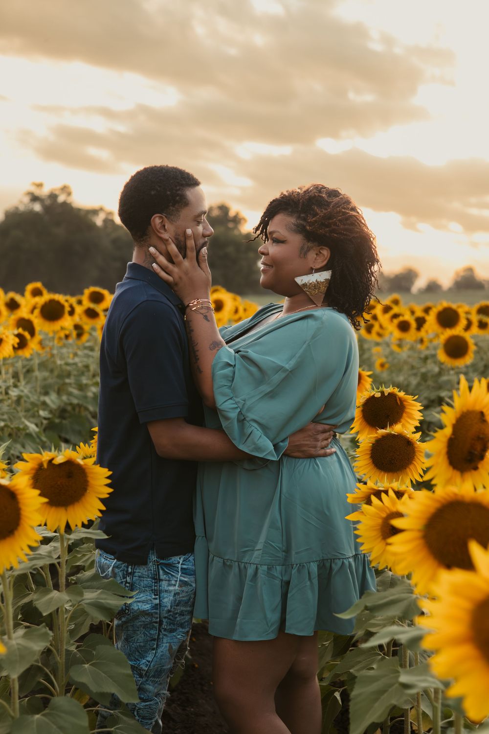A romantic couple embraces in a sunflower field at sunset with golden light illuminating the scenery around them.